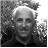 Black and white portrait of a smiling man with short light hair, outdoors with trees and bushes in the background.