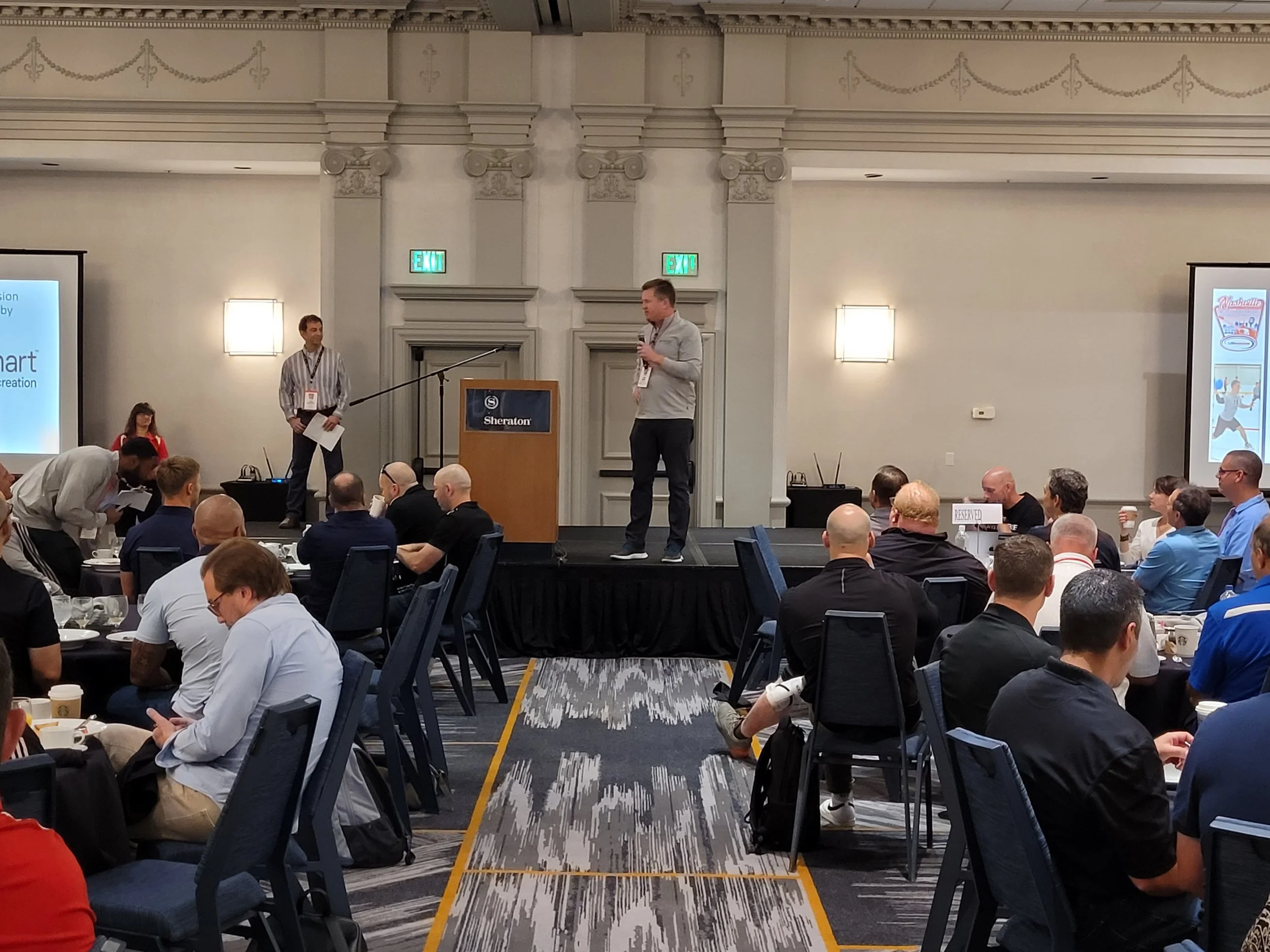 A conference room at a Sheraton hotel filled with attendees sitting at round tables listening to a speaker on stage. The speaker holds a microphone and stands behind a podium displaying the Sheraton logo. There are two screens on either side of the s