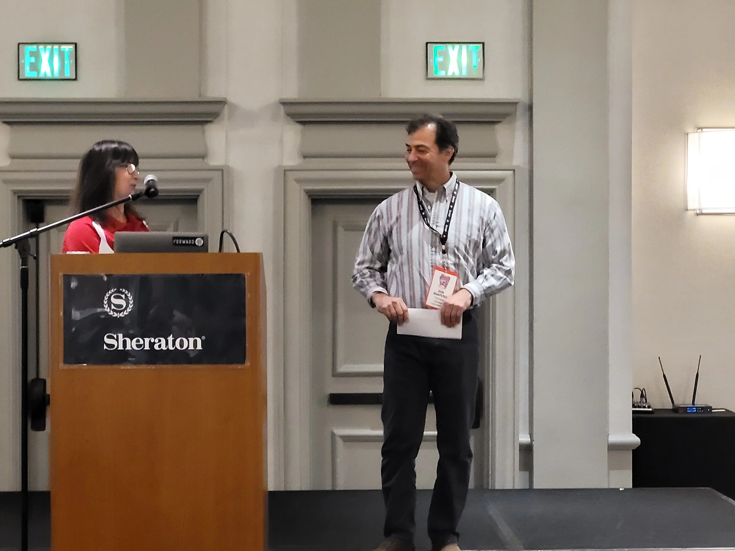 A man in a striped shirt standing on stage, holding a piece of paper and smiling at a woman at a podium with a Sheraton sign. The woman is speaking into a microphone, wearing glasses and a red shirt. There are two illuminated EXIT signs on the wall b