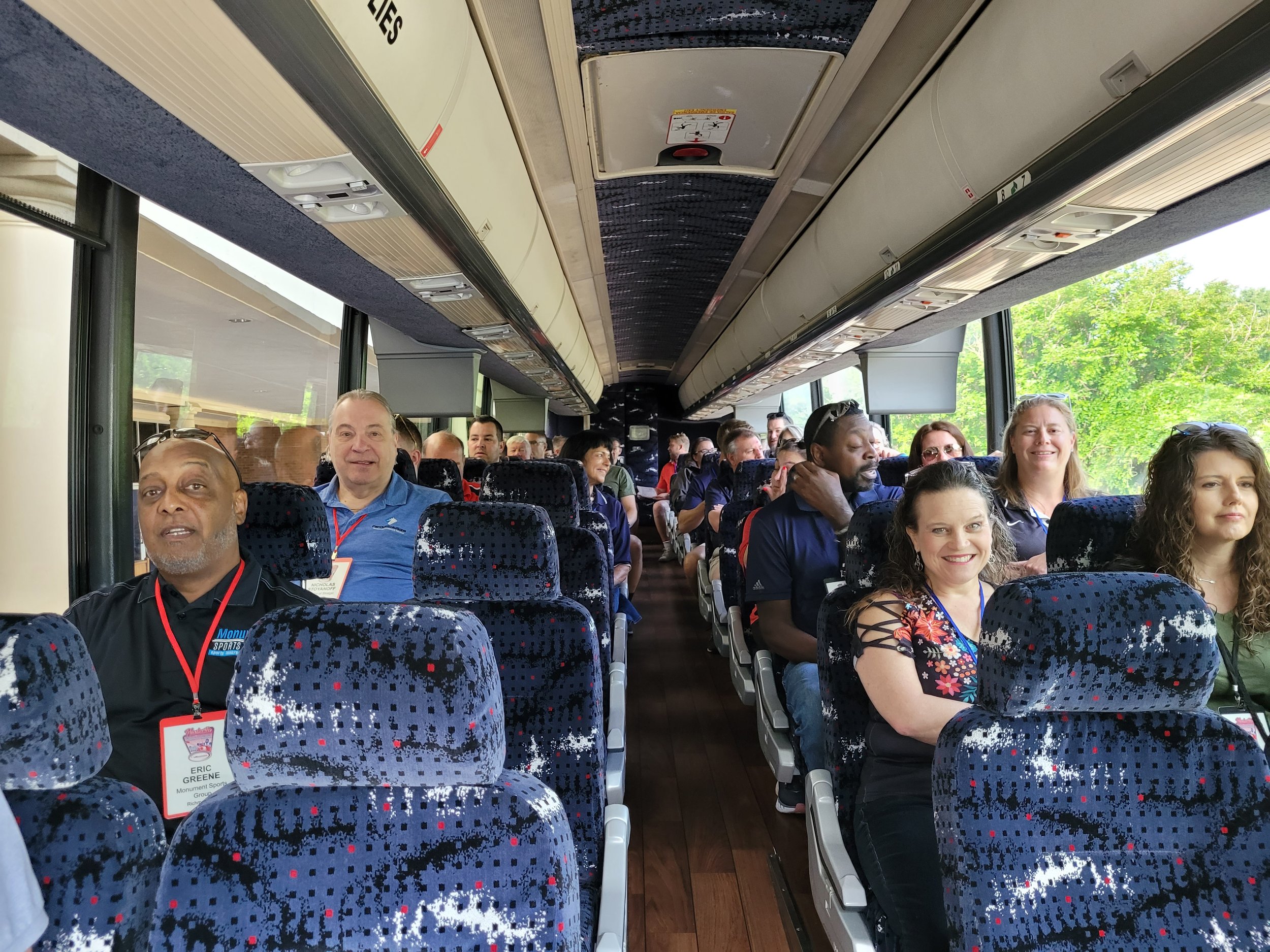 Inside a bus filled with diverse passengers seated and facing forward, with large windows showing green trees outside, and overhead luggage compartments.