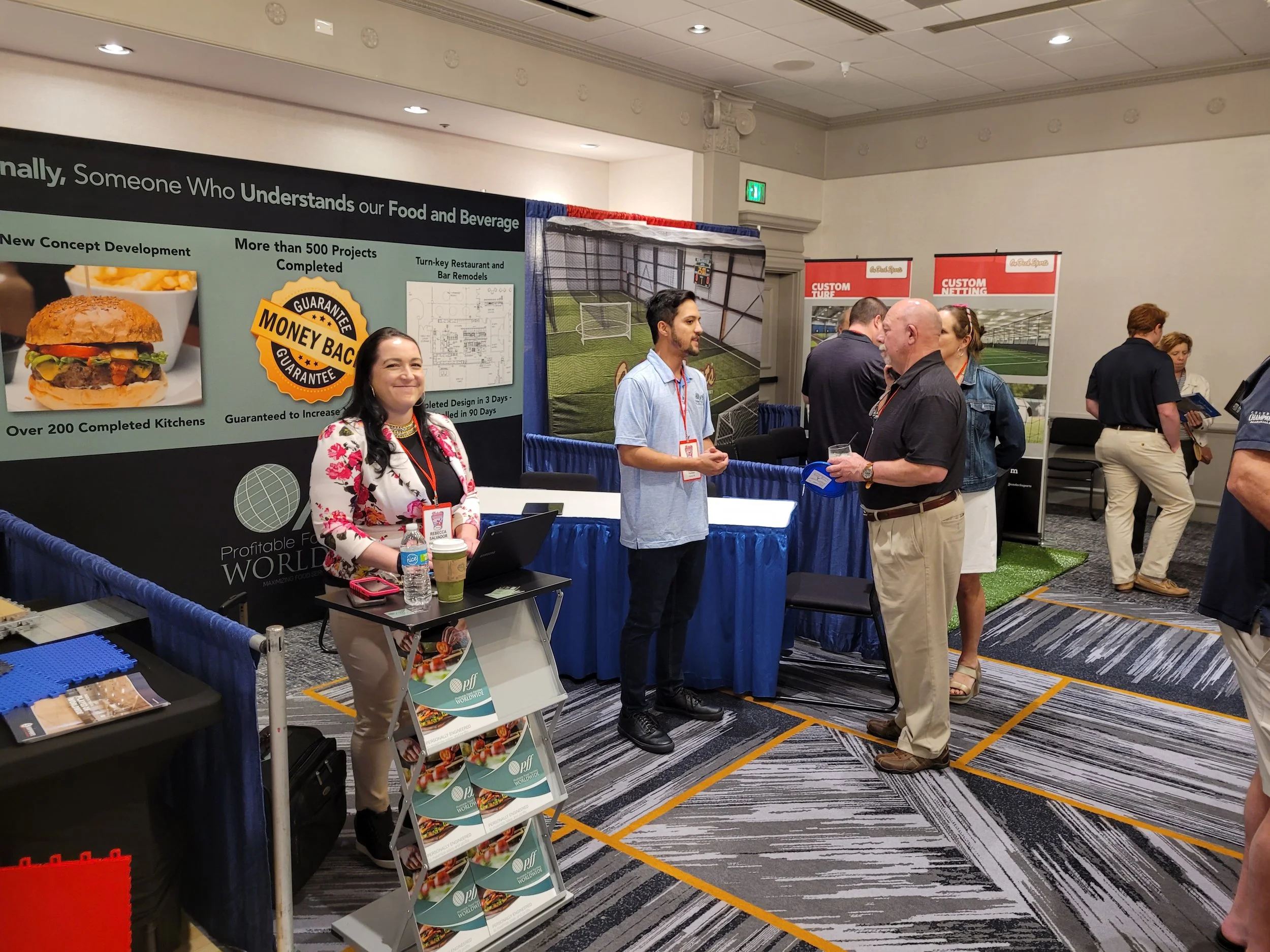 At a trade show, a woman stands behind a booth with a large display promoting food and beverage development, and a man is talking to another attendee. People are browsing and engaging with different booths in the room.