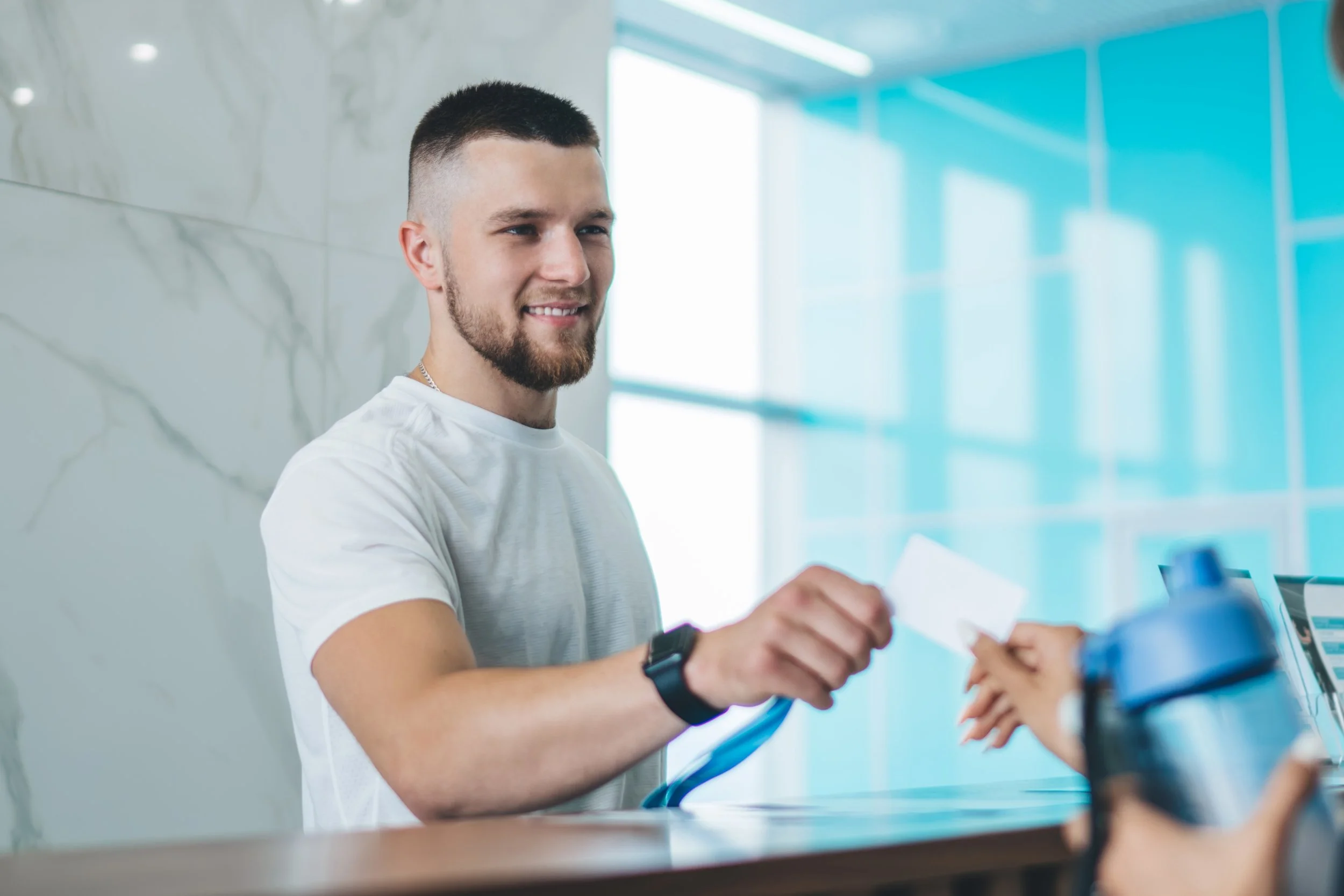 A young man with short dark hair and a beard smiling while handing over a piece of paper to another person at a counter in a bright, modern setting with blue-tinted glass walls.