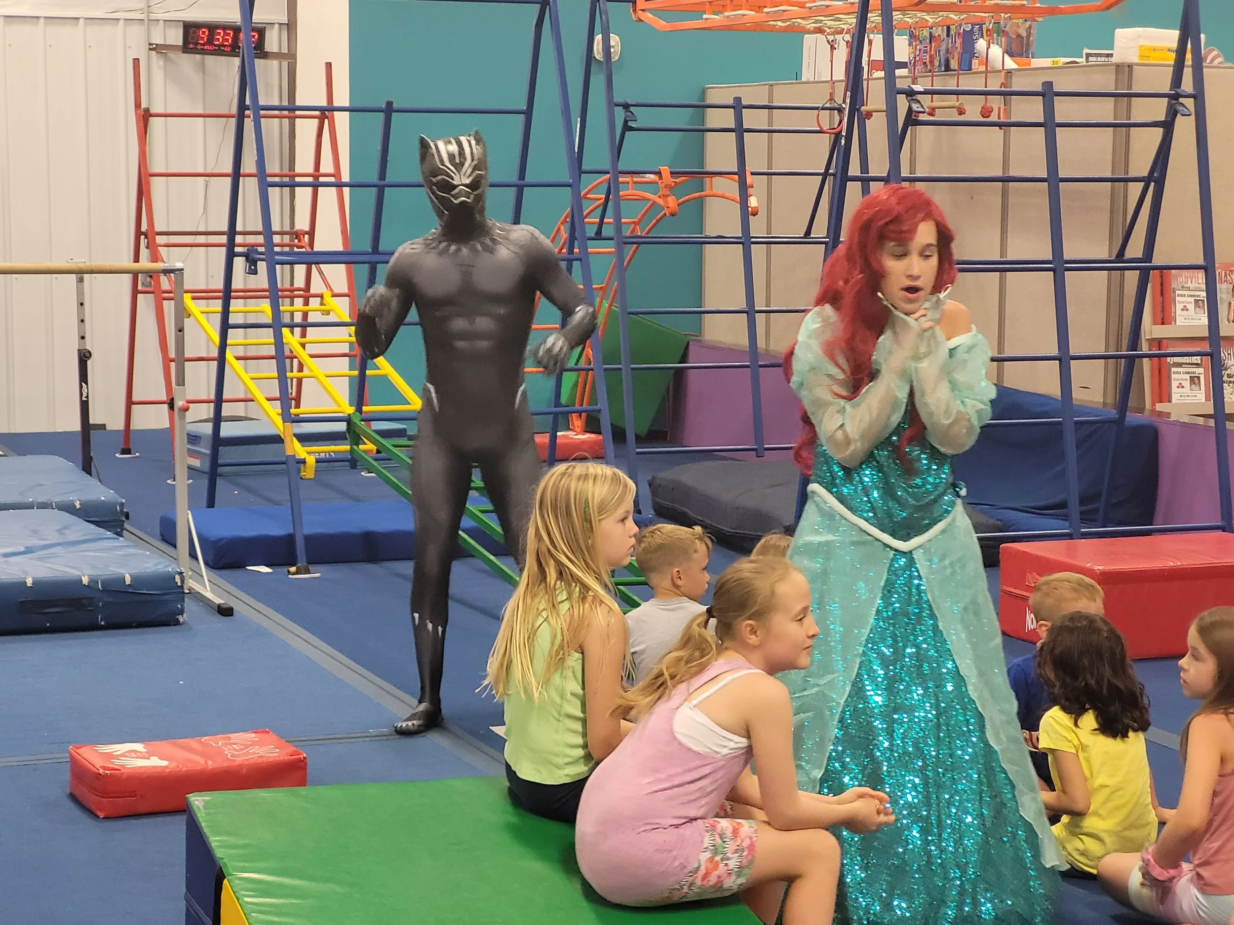 A group of children seated on the floor watching a woman dressed as Elsa from Frozen, with red hair and a sparkling blue dress. Behind them is a colorful indoor play area with climbing structures and a tarp ceiling.
