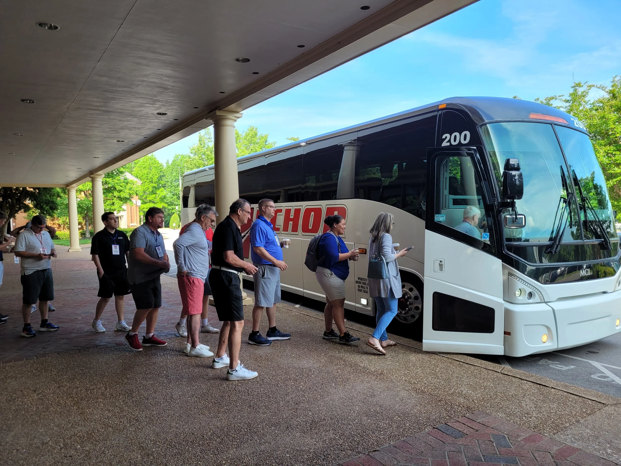 People waiting in line to board a white tour bus parked under a covered area with columns, surrounded by green trees and a blue sky.
