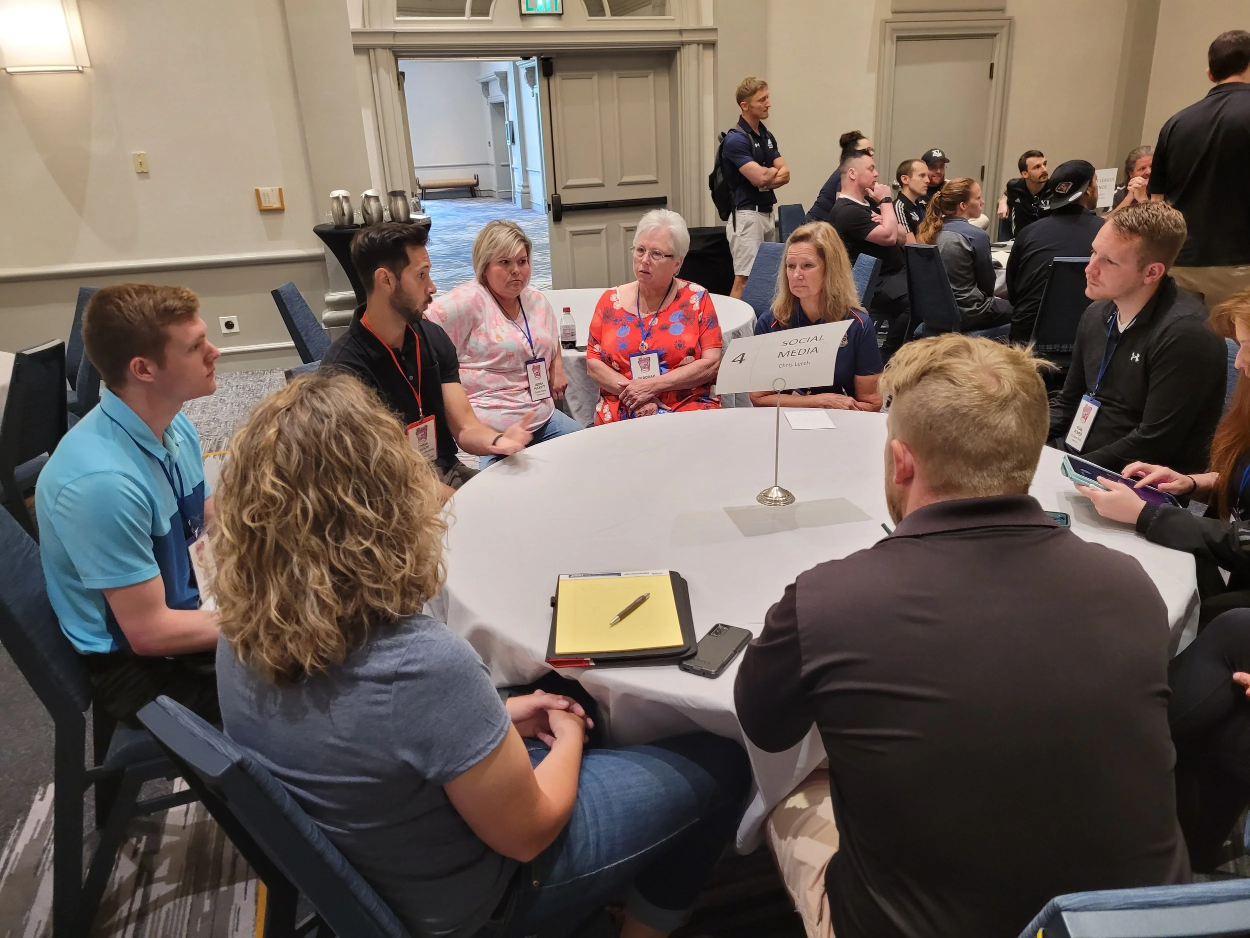 A group of people seated around a round table in a conference room, engaged in a discussion. Some individuals are wearing name tags, and a sign on the table reads 'Social Media.' There are more people standing in the background, and the room has neut