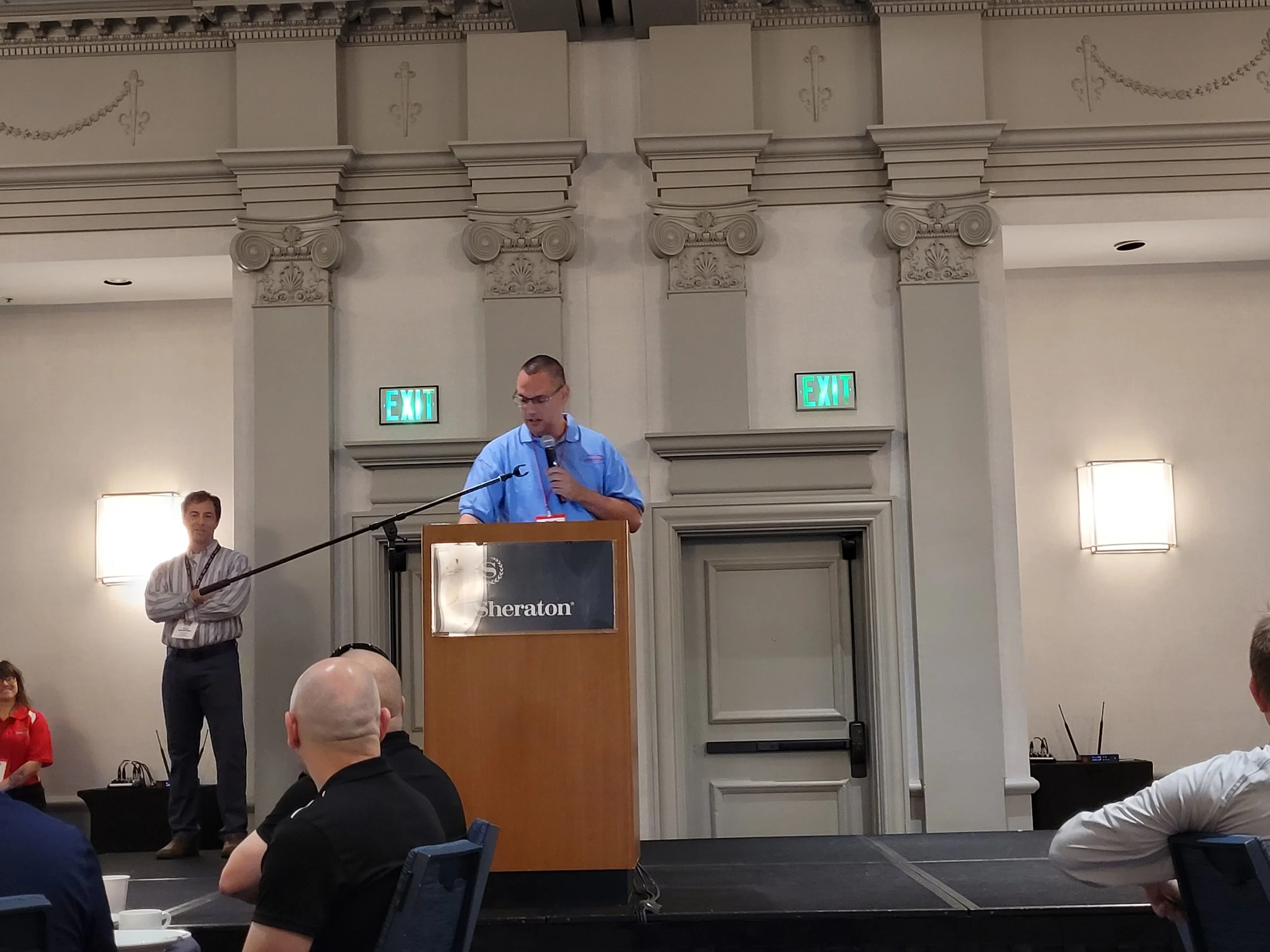 A man in a blue shirt is standing at a podium with a microphone, speaking to an audience in a large, elegant conference room with high ceilings, ornate moldings, and two illuminated exit signs. There are people seated and listening, and a person in a