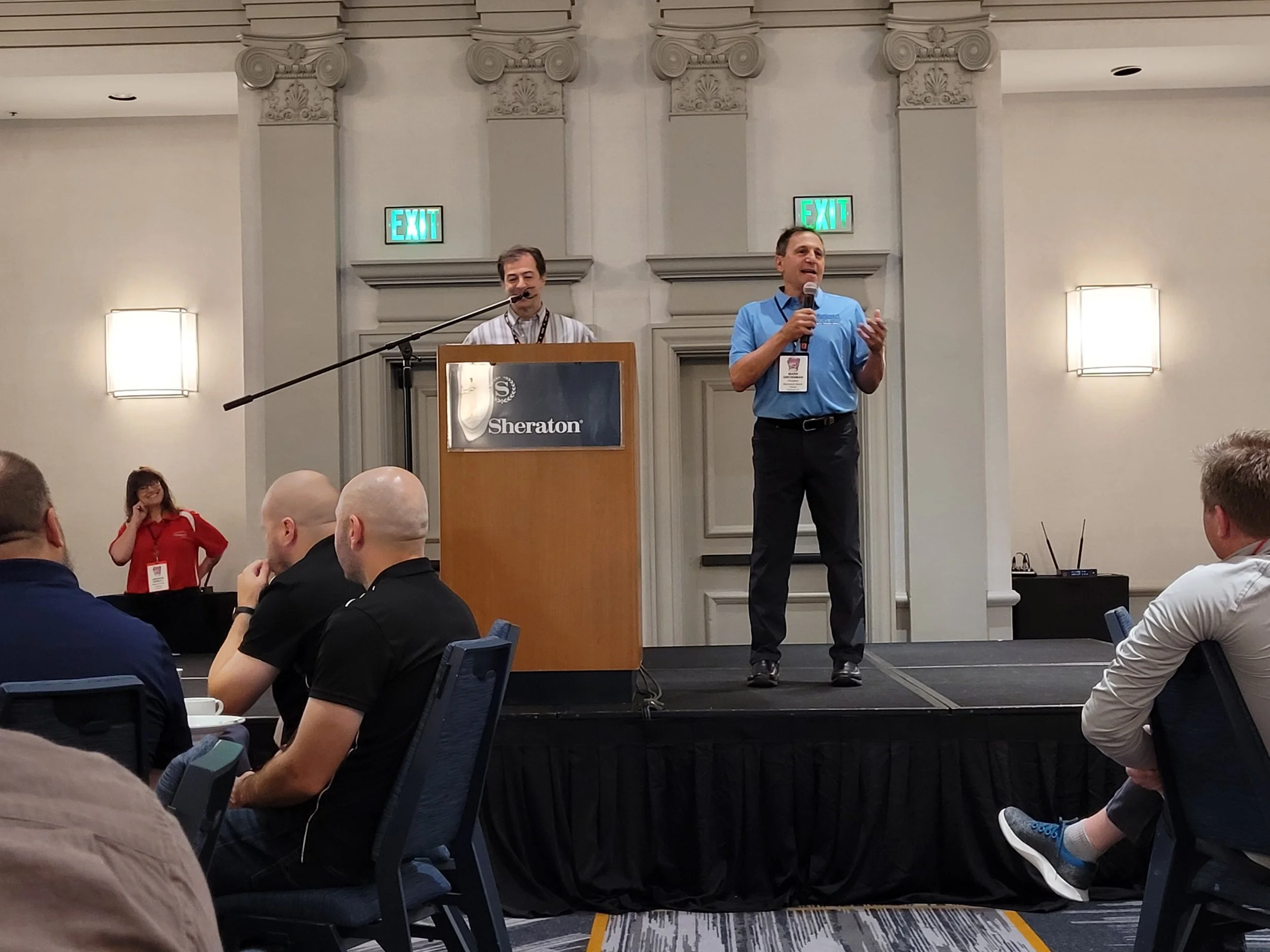 A man in a blue polo shirt is speaking into a microphone while standing on a small stage. Behind him, another man is smiling at a podium with a Sheraton sign. Several people are seated at tables, listening to the presentation in a conference room wit