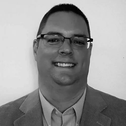 A Black and white photo of a man wearing glasses, a suit, and a collared shirt, smiling while facing the camera.