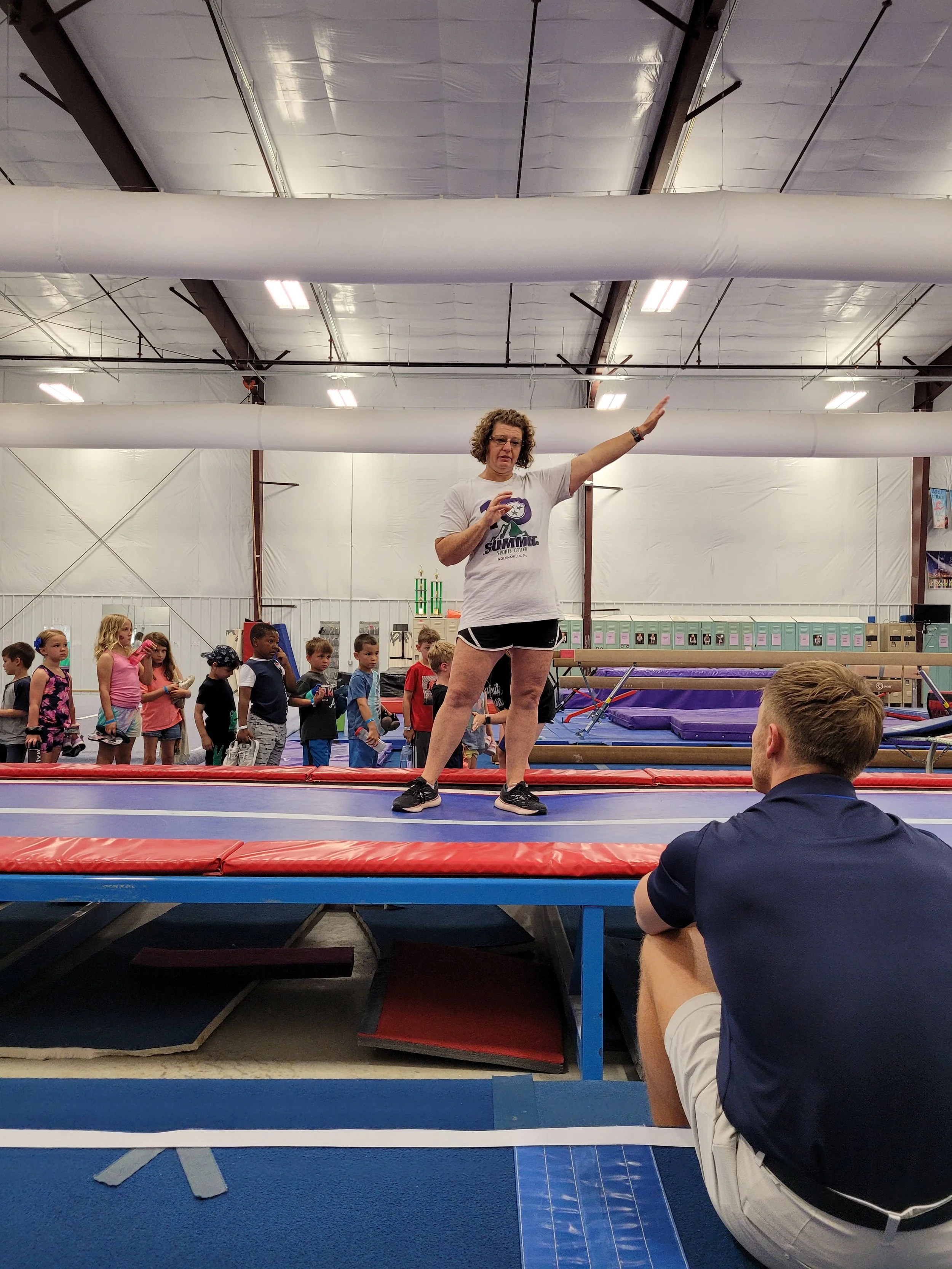 A woman standing on a trampoline in a gymnastics gym, demonstrating a pose or movement, with children and gym equipment in the background.