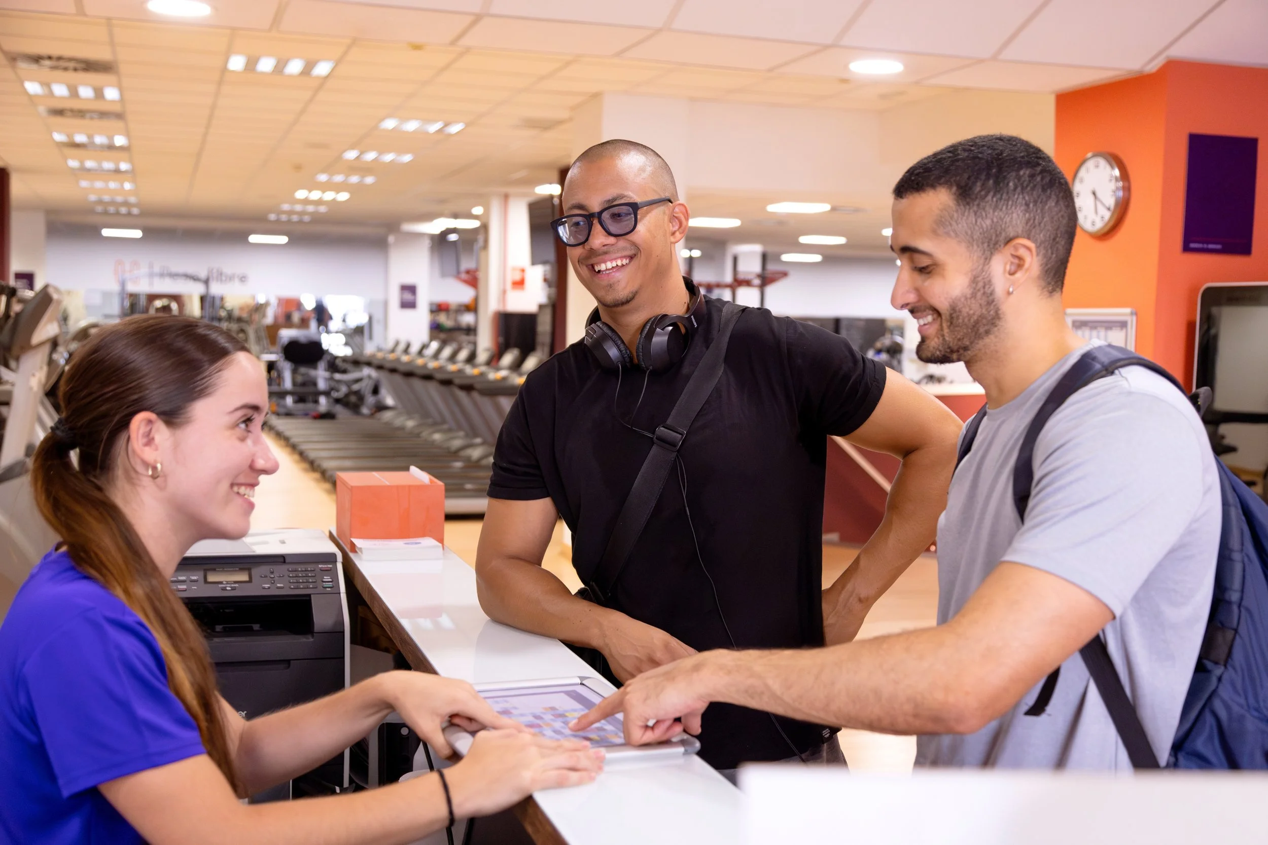 Two men and a woman are at a check-in counter at an airport, smiling and engaging with each other. The woman wears a blue shirt, the man in the middle has glasses and headphones around his neck, and the other man has a backpack.