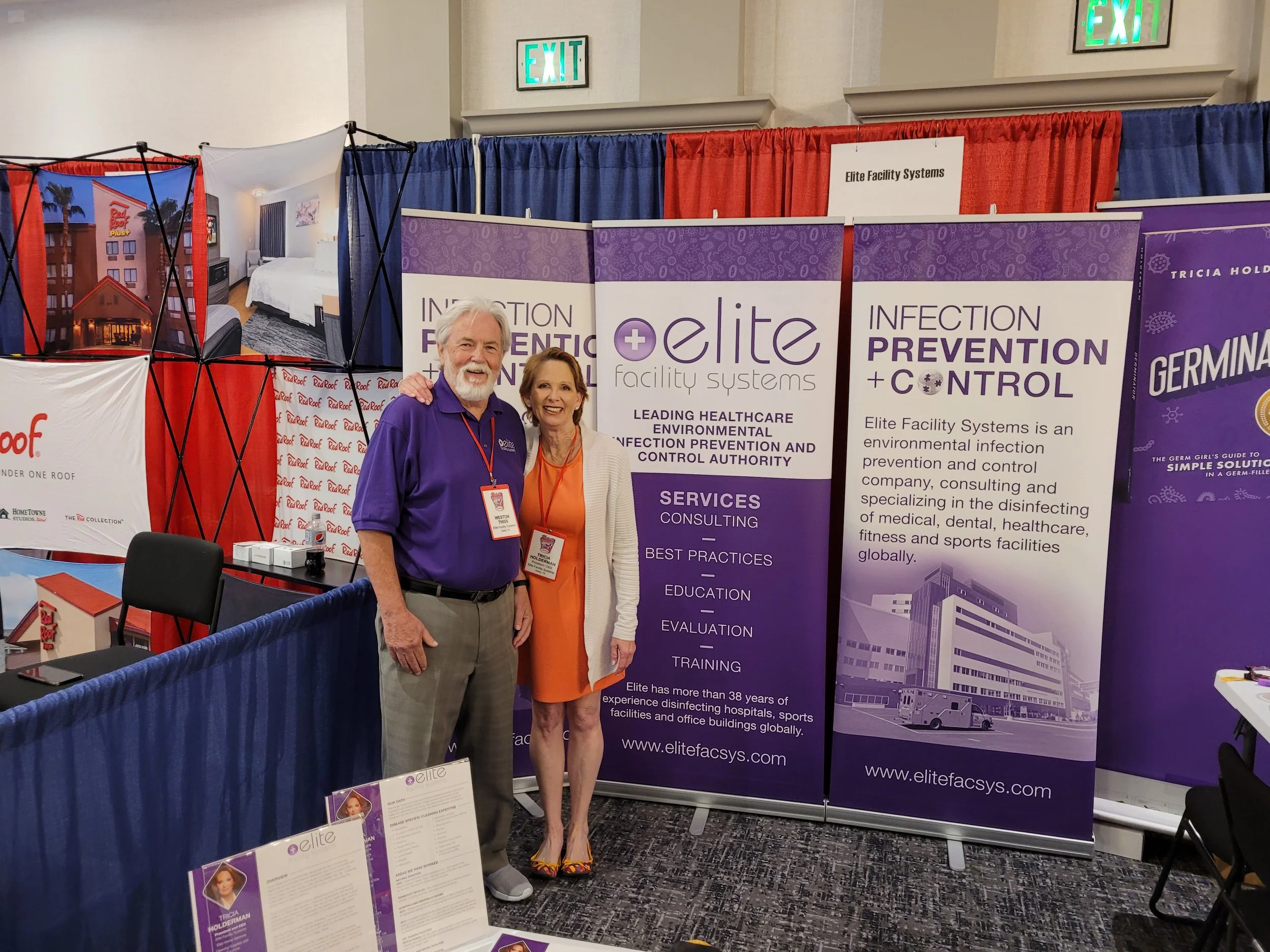 Two people standing in front of a booth display at a conference or trade show. The booth features large purple banners with white and purple text about Elite Facility Systems, a healthcare infection prevention company. The man has gray hair and beard