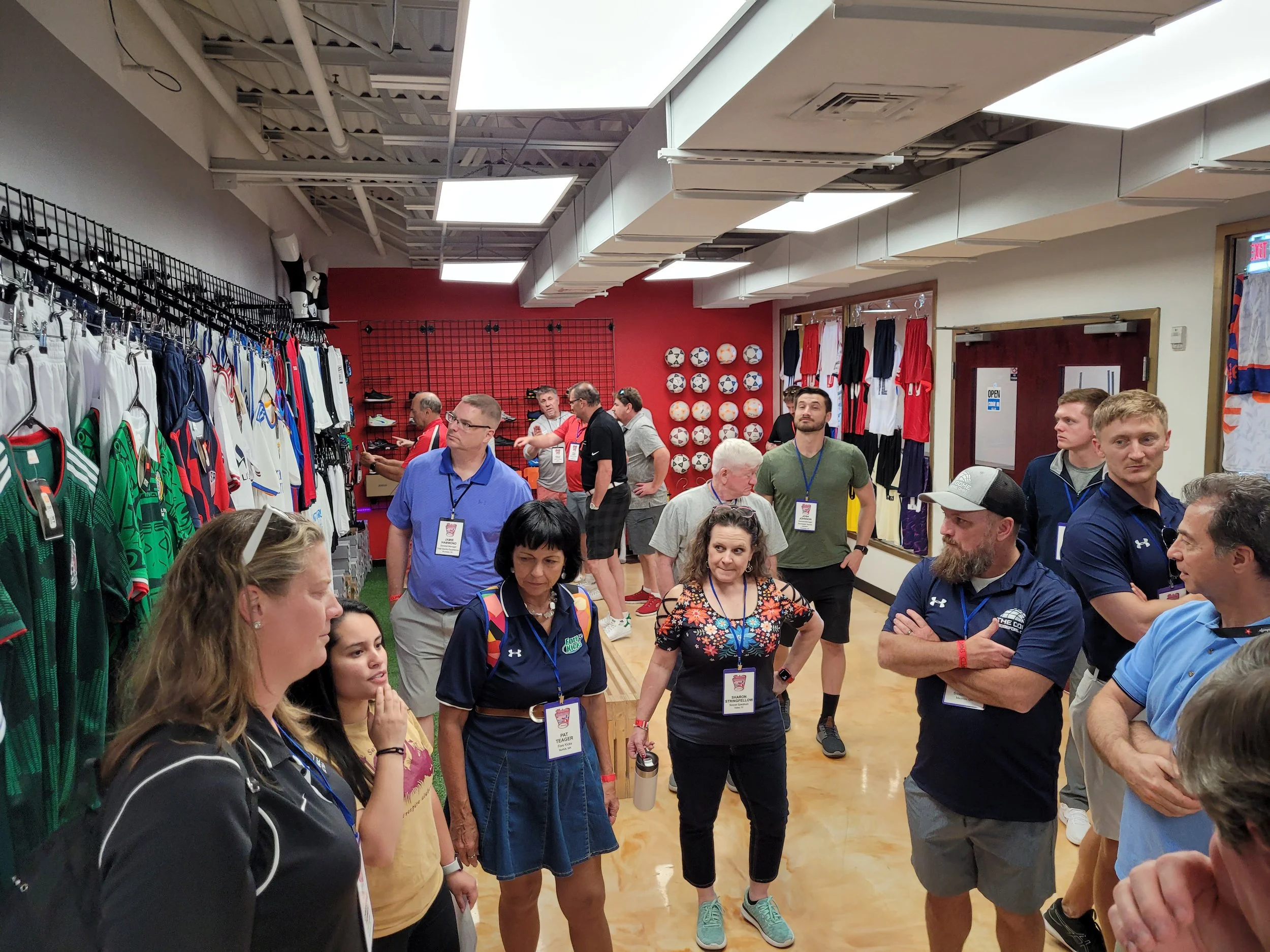 A group of people gathered indoors, some with name tags, surrounded by sports apparel and memorabilia, with a red wall displaying soccer balls in the background, indicating a social or networking event related to sports.