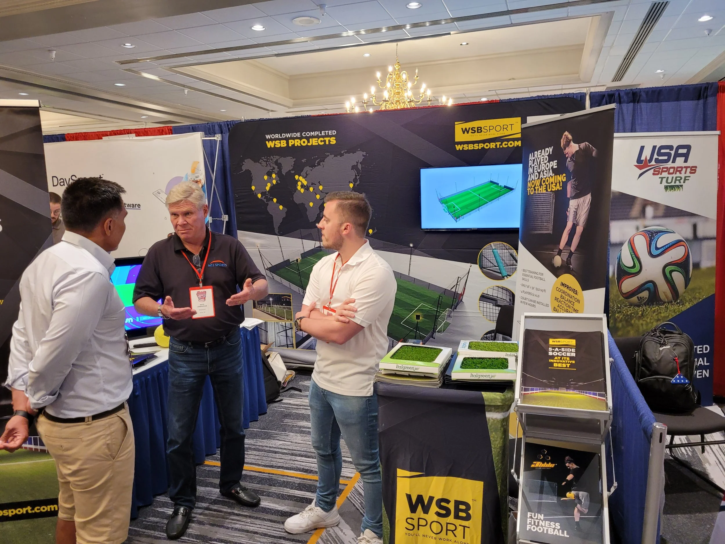 Three men are standing and talking at a sports equipment booth at an indoor trade show. The booth displays a large banner with a world map and the text 'Worldwide Completed WSB Projects' and 'WSSPORT.COM', with imagery of a football field on a screen