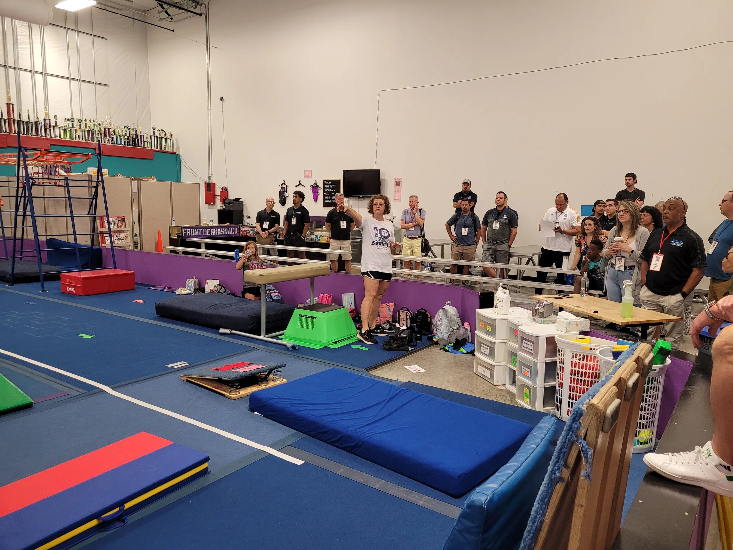 People gathered in an indoor gym or activity center with gymnastic mats, weight benches, and a climbing frame. Some individuals are standing and listening to a person speaking.