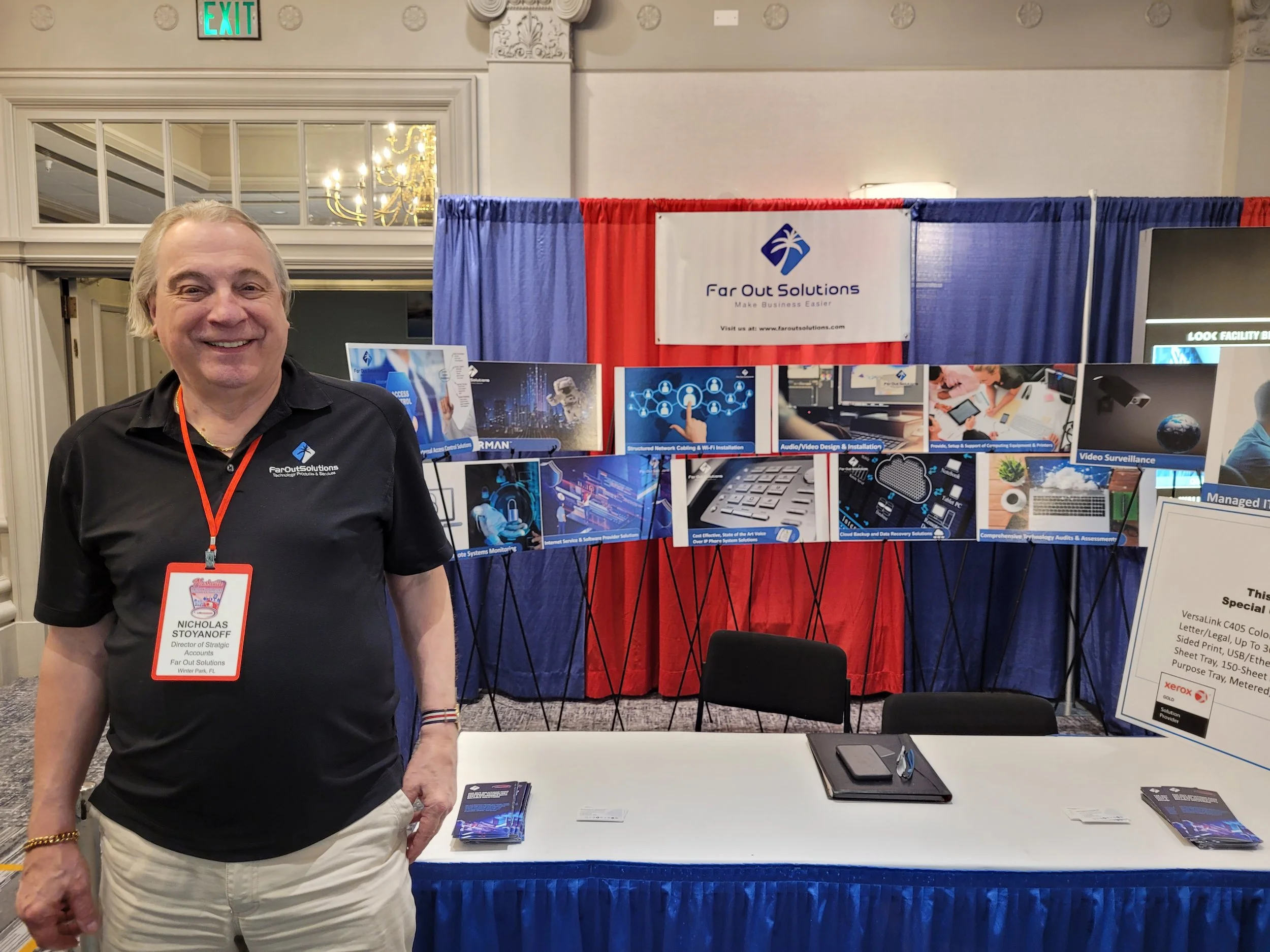 A man wearing a black shirt with a name tag stands in front of a booth at a conference or trade show. The booth has a red, white, and blue curtain backdrop and displays brochures, posters, and a laptop. The man is smiling and looking at the camera, a