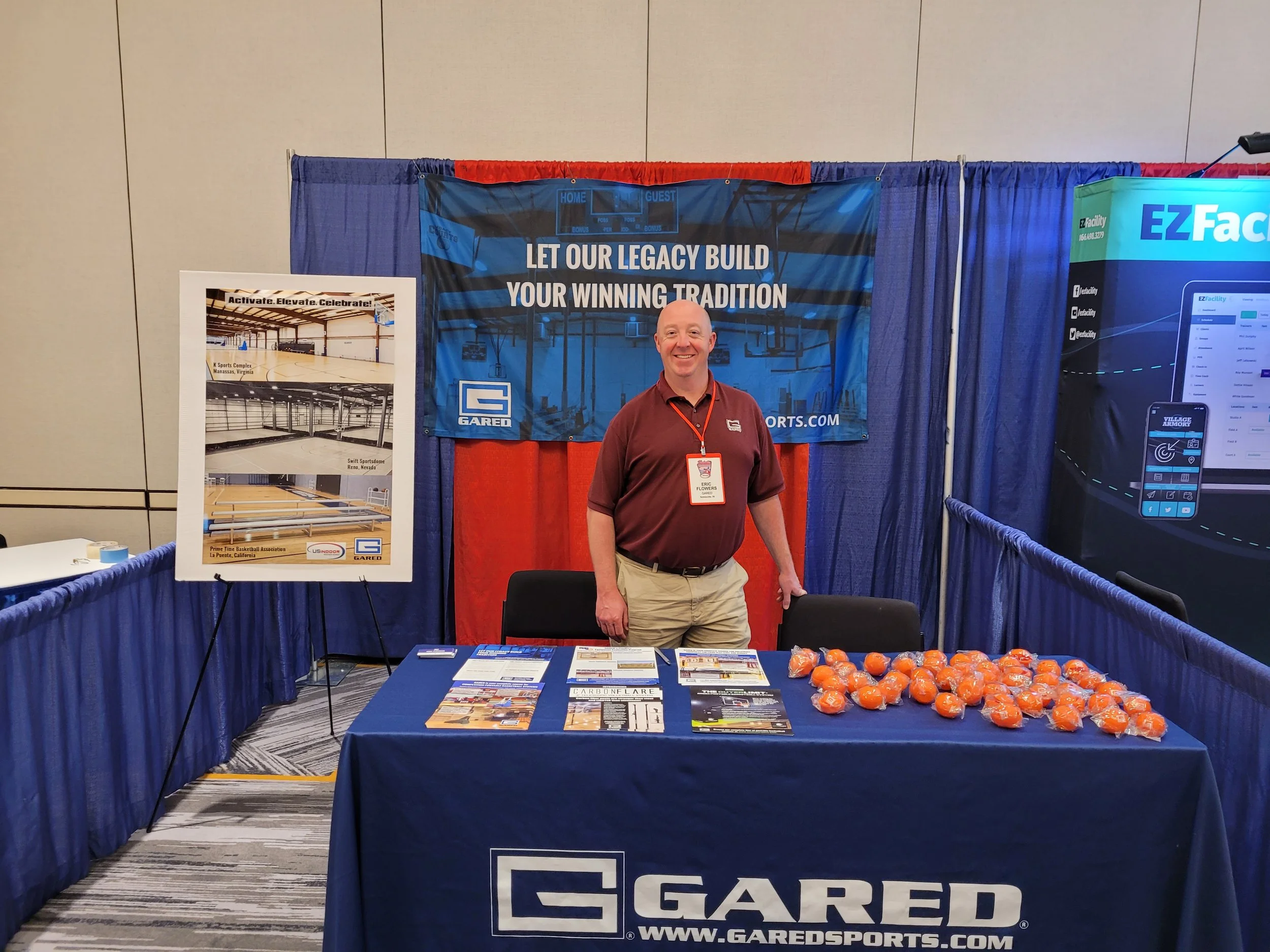 A man standing at a Gared Sports booth with a blue backdrop that reads "Let our legacy build your winning tradition." The booth features a table with brochures, a cluster of orange stress balls, and a sign showcasing the Swift Sports Arena in Reno, N