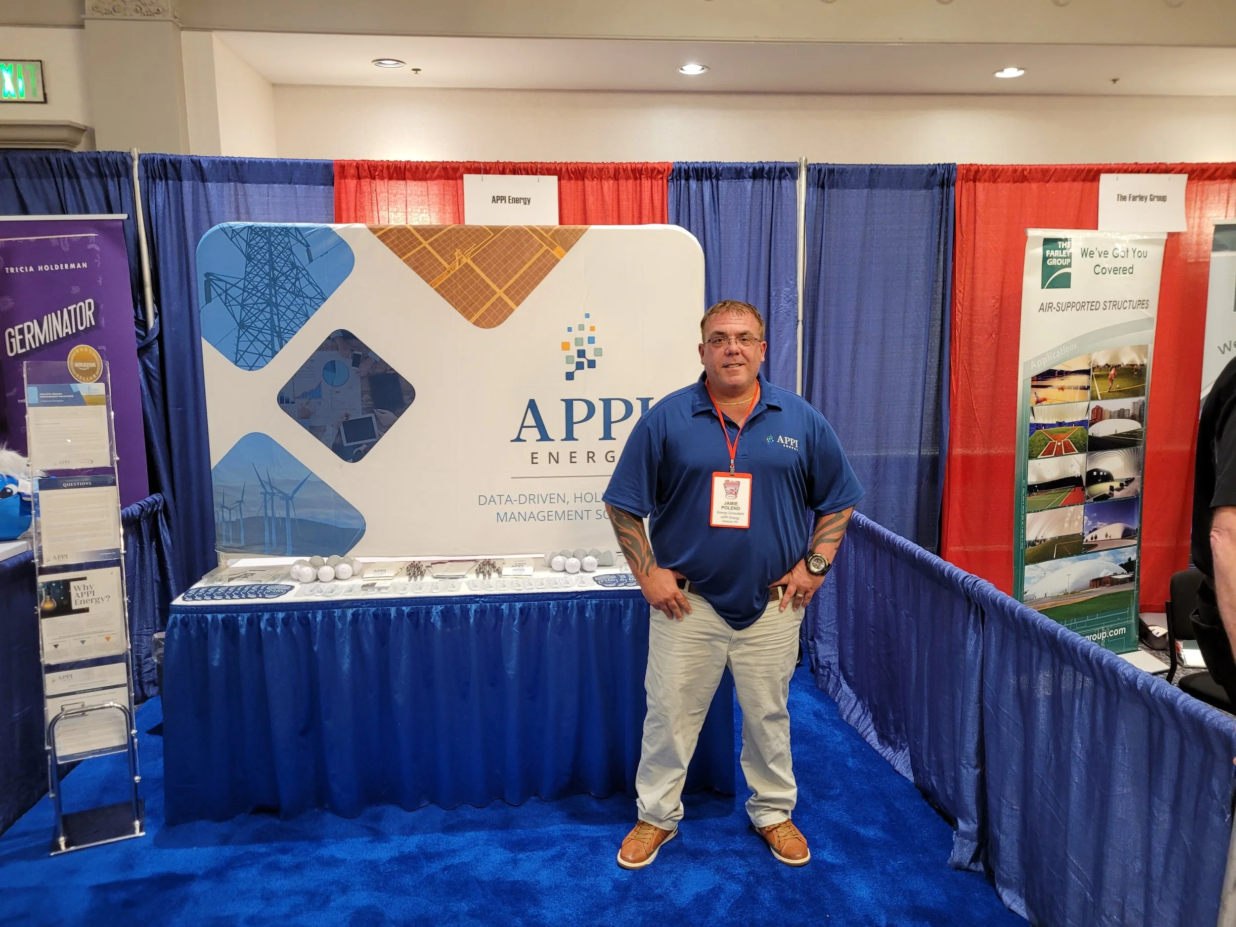 A man standing in front of a booth at a conference or expo. The booth has a large sign with the logo and name of APP Energy, featuring wind turbines and solar panels. The man is wearing a blue polo shirt with APP Energy and a name badge that says Jam