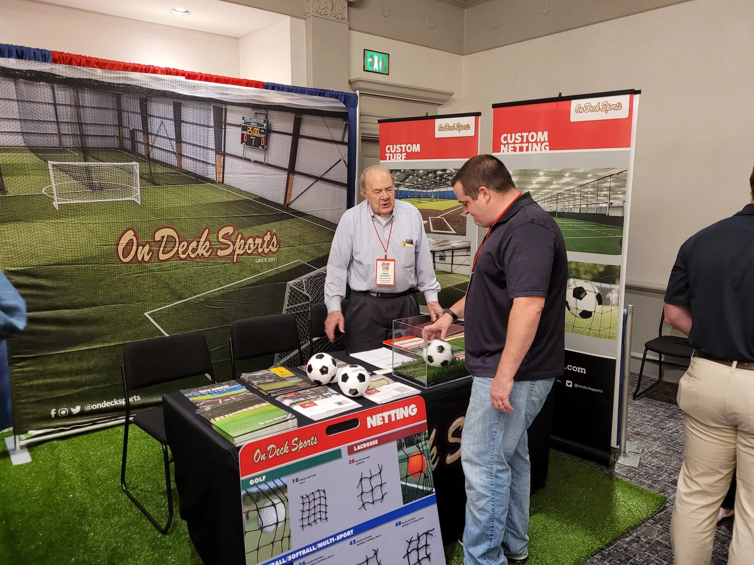 At an indoor sports expo, a booth showcases different types of sports netting and related equipment, with posters and display items including soccer balls and nets, and two men engaging in conversation.