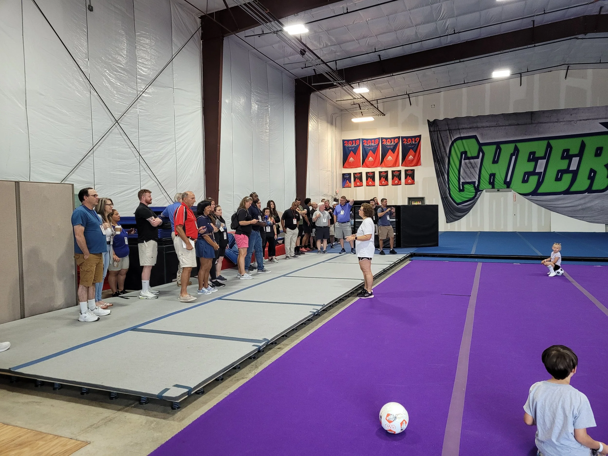 A group of people standing in line in an indoor gymnasium, waiting for their turn to participate in a cheerleading or gymnastics event. There is a large banner with the word 'CHEER' and some championship banners on the wall. A few children are sittin