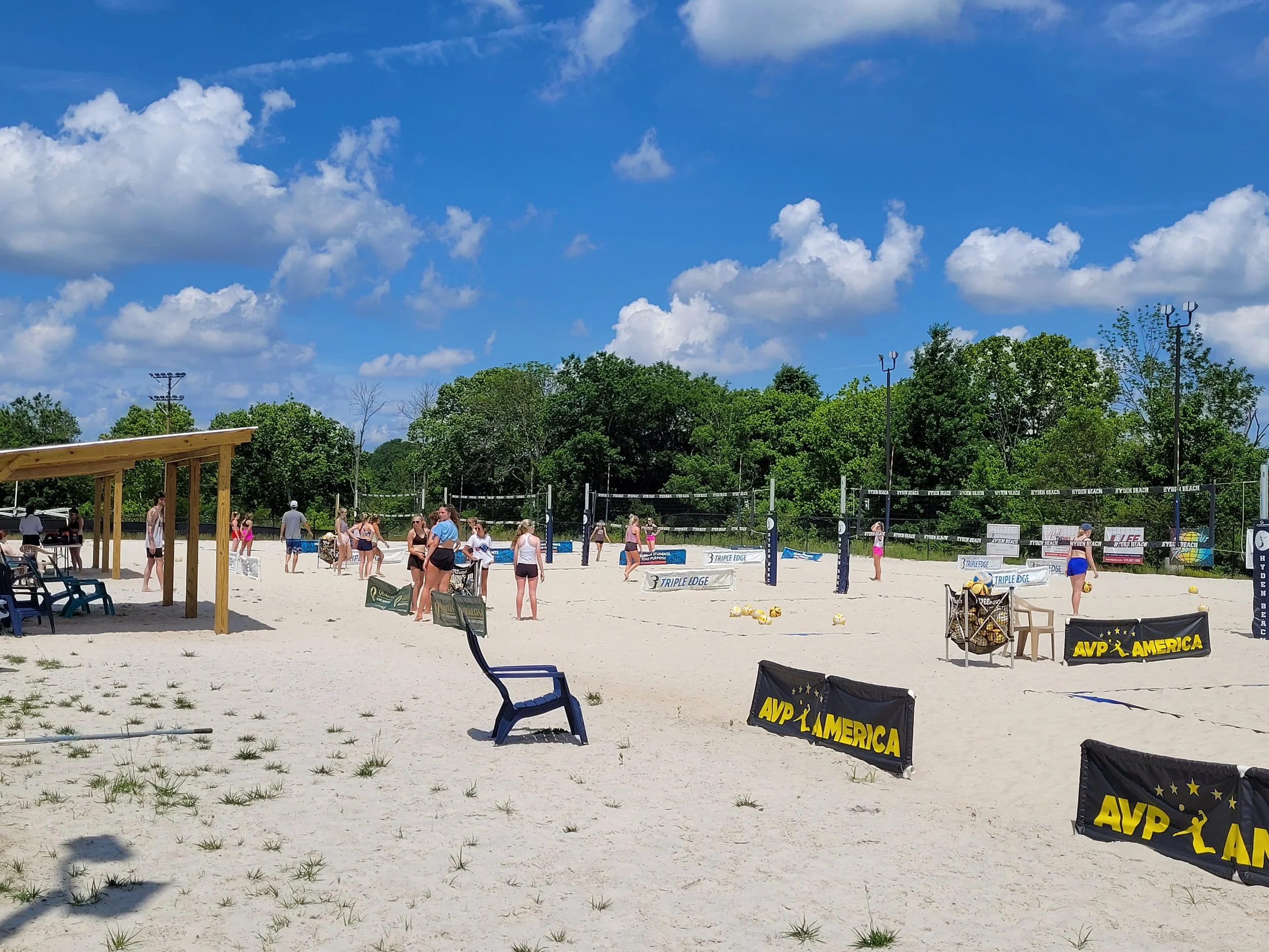 People playing volleyball on a sandy beach court under a partly cloudy sky, surrounded by green trees and sporting banners.