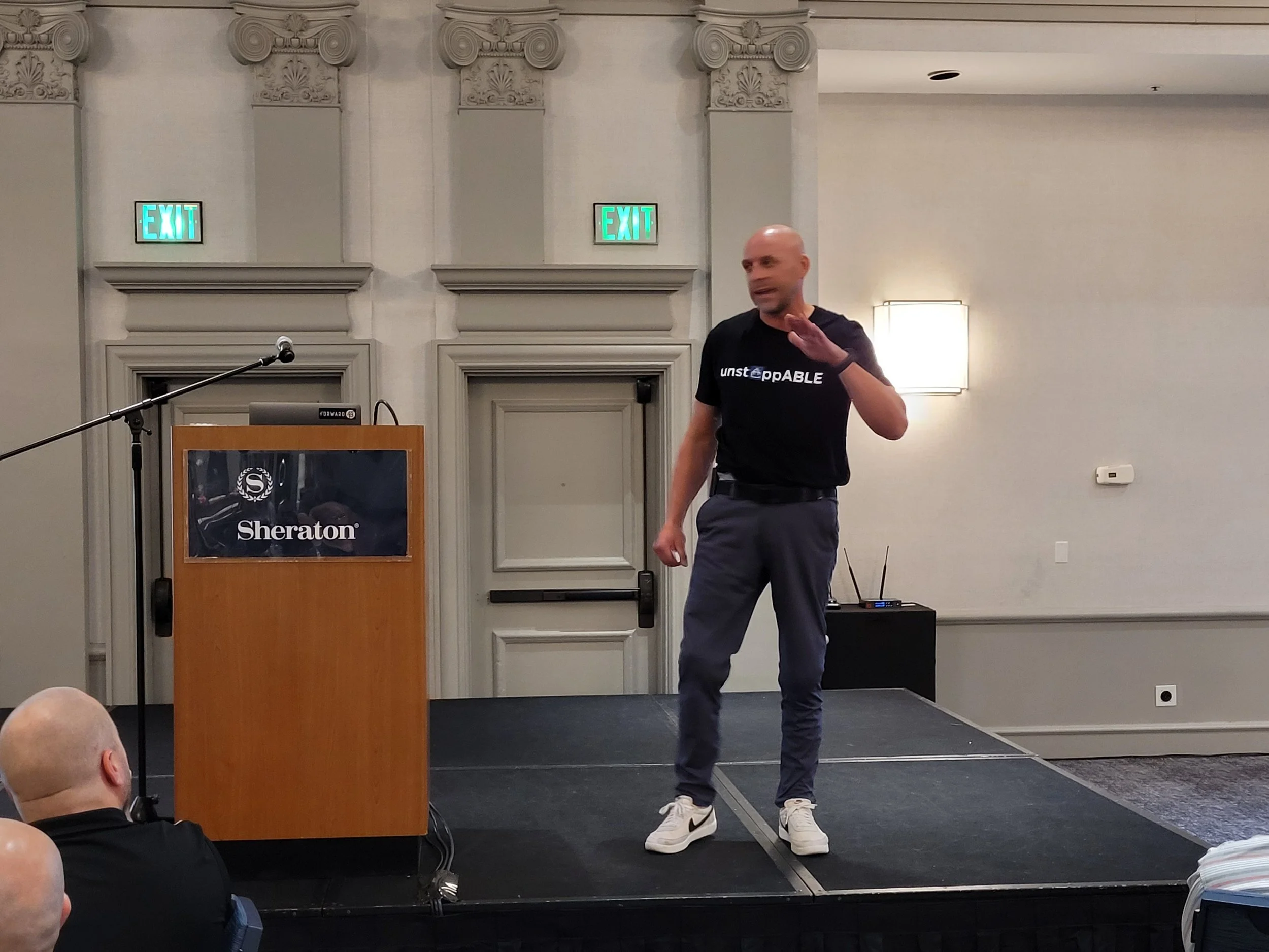 A man wearing a black T-shirt with 'unstoppable' printed on it is standing and gesturing with his right hand in a conference room. There is a wooden podium with the Sheraton logo, a microphone, and a laptop. The room has beige walls, decorative ceili