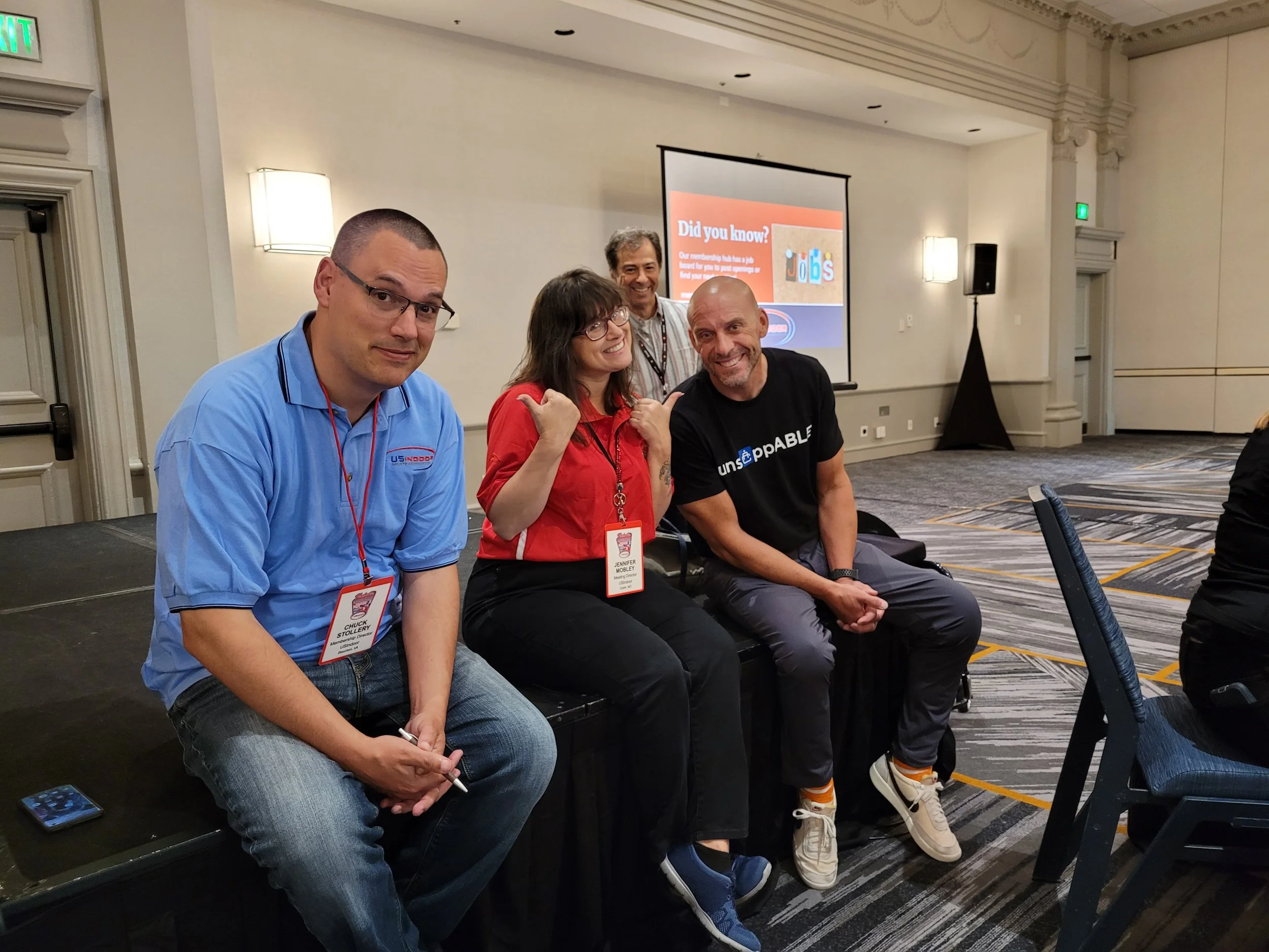 Four people sitting on a bench at an indoor event, smiling at the camera. There is a large screen behind them with a presentation slide that says 'Did you know?'. The individuals are wearing casual clothes and name tags.