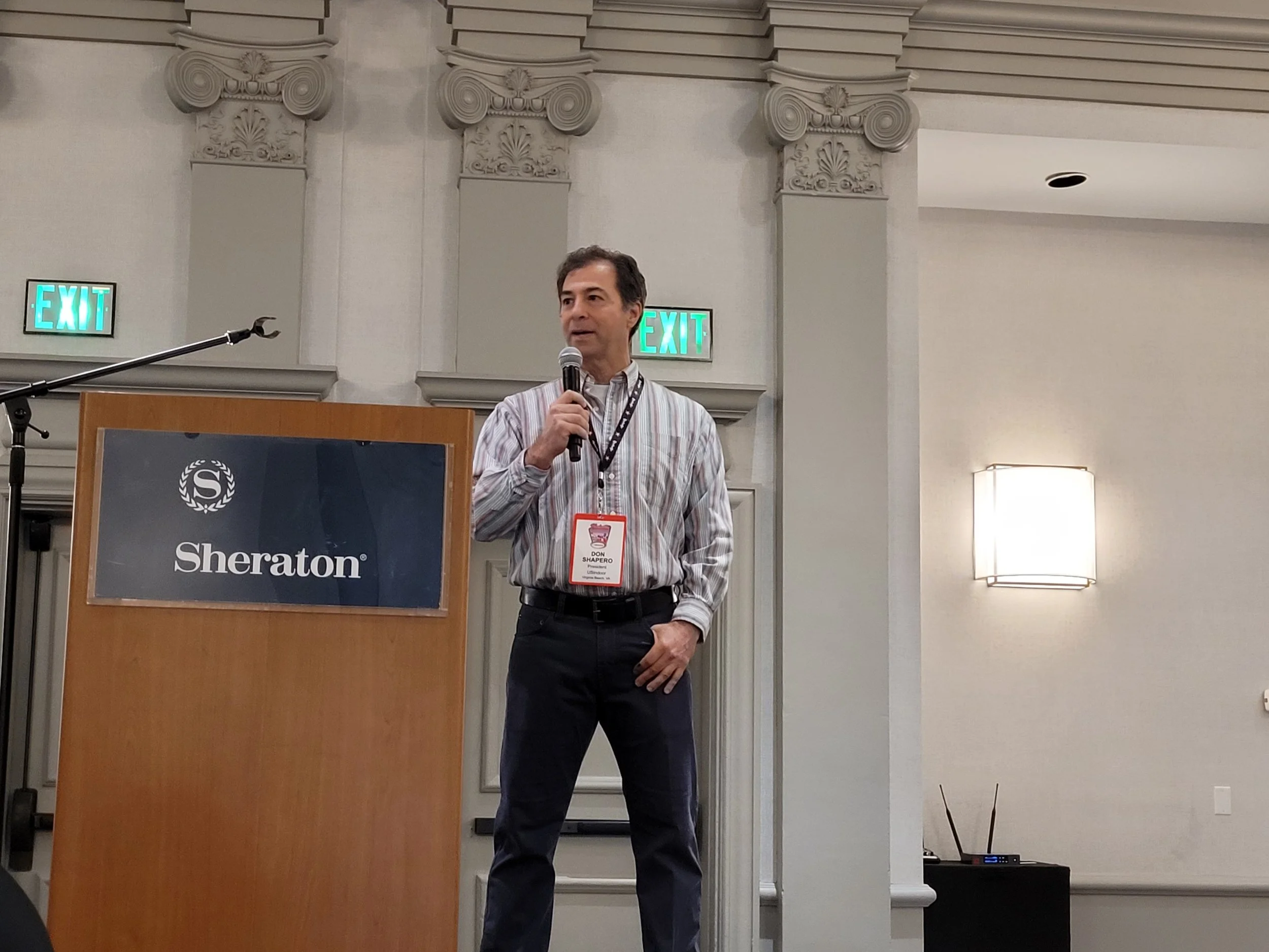 A man in a striped shirt holding a microphone and speaking at a podium with a Sheraton logo, standing indoors near a wall with decorative columns and two illuminated exit signs.