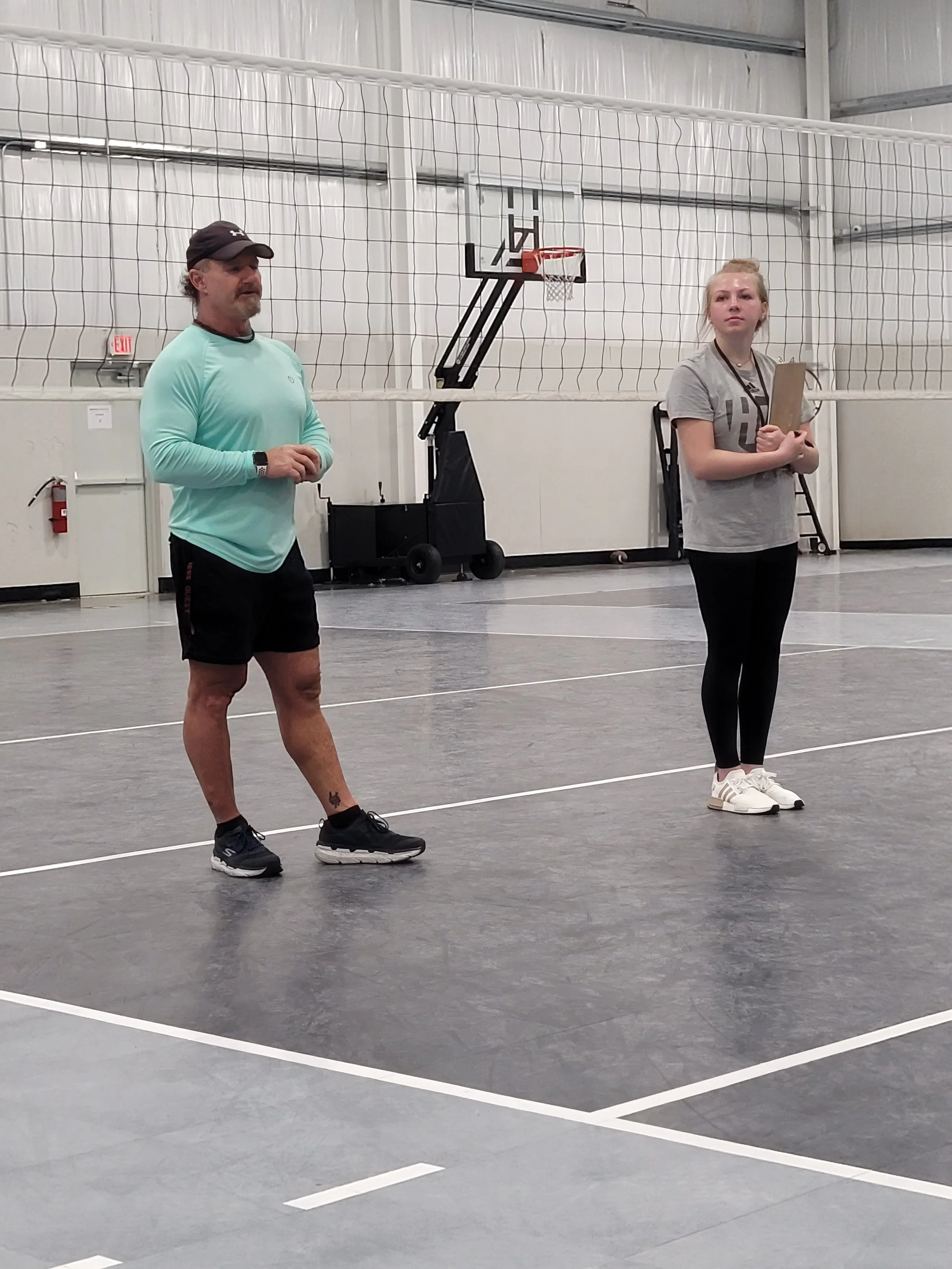 Two people standing on a gymnasium basketball court, separated by a volleyball net, with a basketball hoop and white walls in the background.