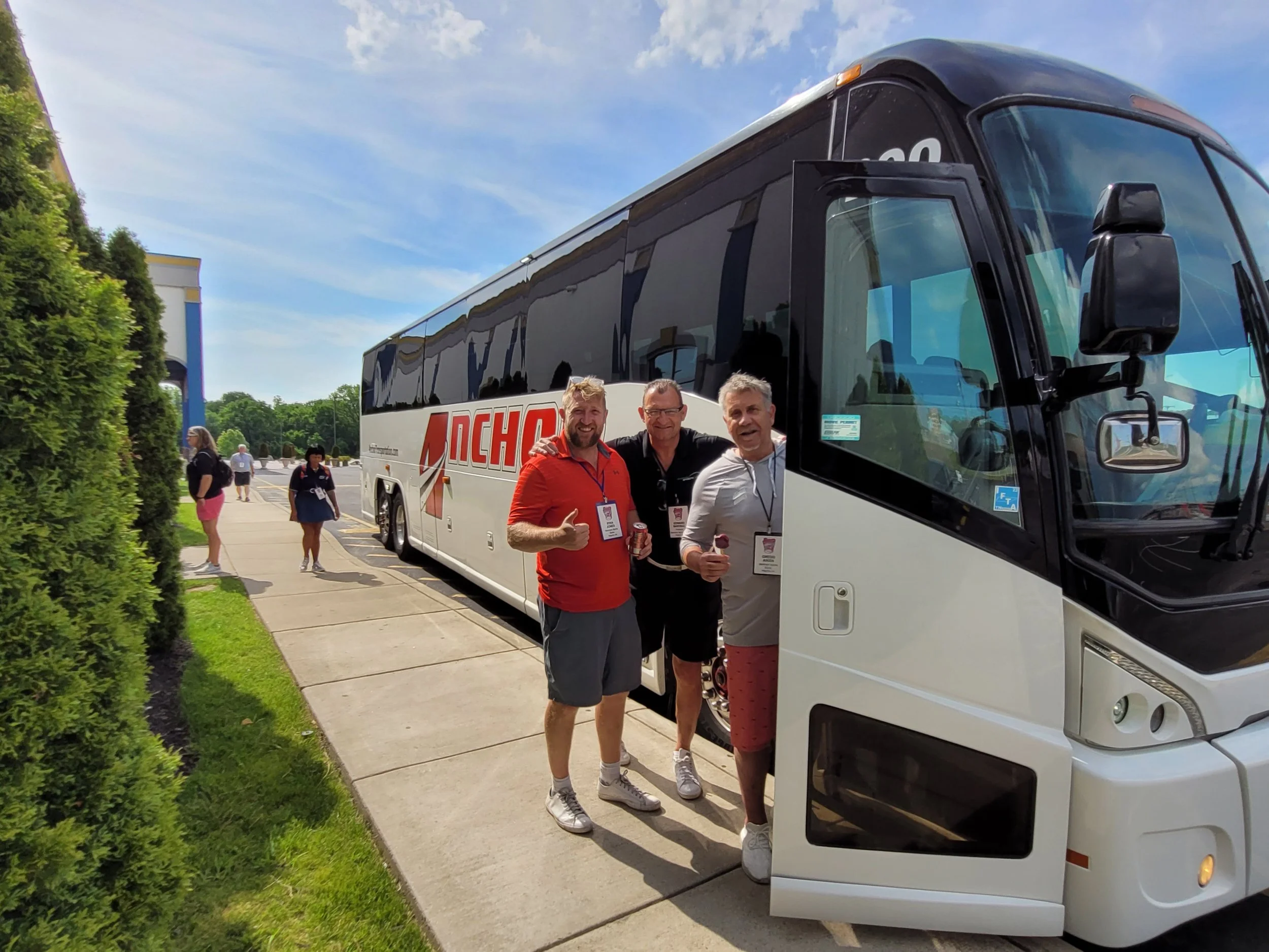 Three men standing in front of a white tour bus, smiling and giving thumbs up. The bus has the word 'AERO' partially visible on its side. Several people are walking in the background on a sidewalk next to a hedge, under a partly cloudy sky.