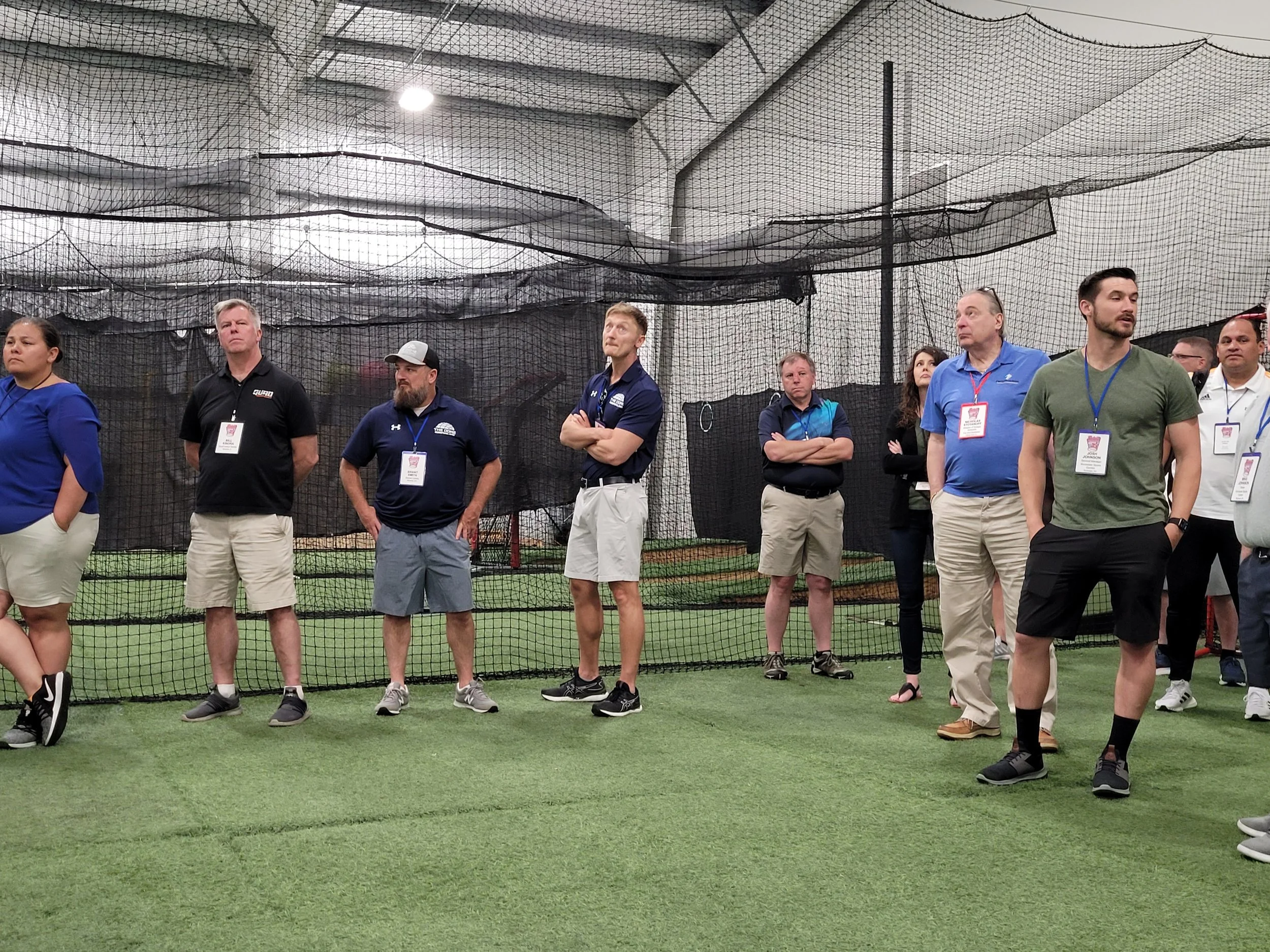 Group of people standing inside an indoor sports facility with a netted ceiling and walls, some wearing badges around their necks.