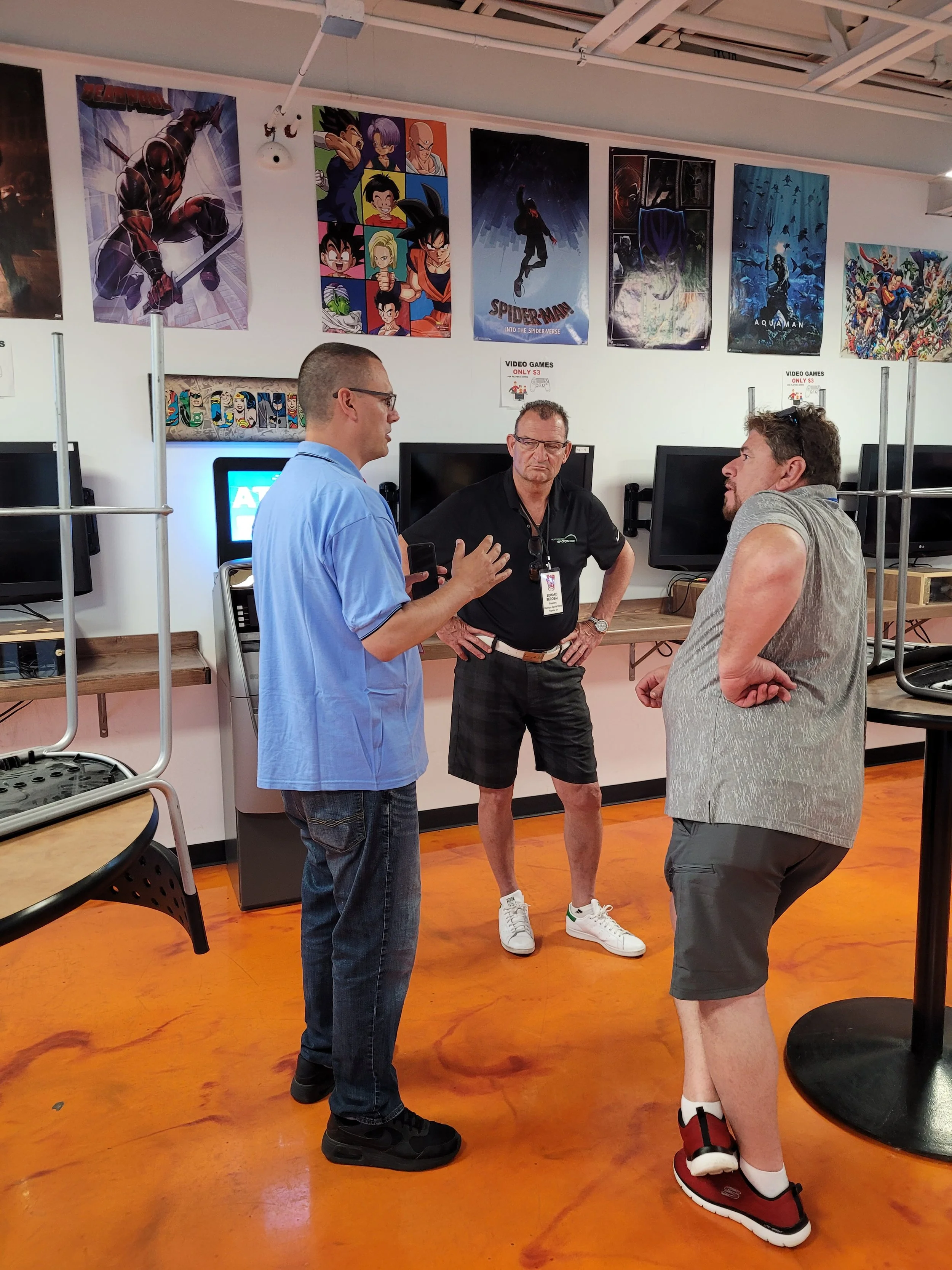 Three men are standing and conversing in a room with many posters of animated characters on the wall. Two of the men are wearing glasses, and all three are dressed in casual clothing. The room appears to be a gaming or office space with computers and