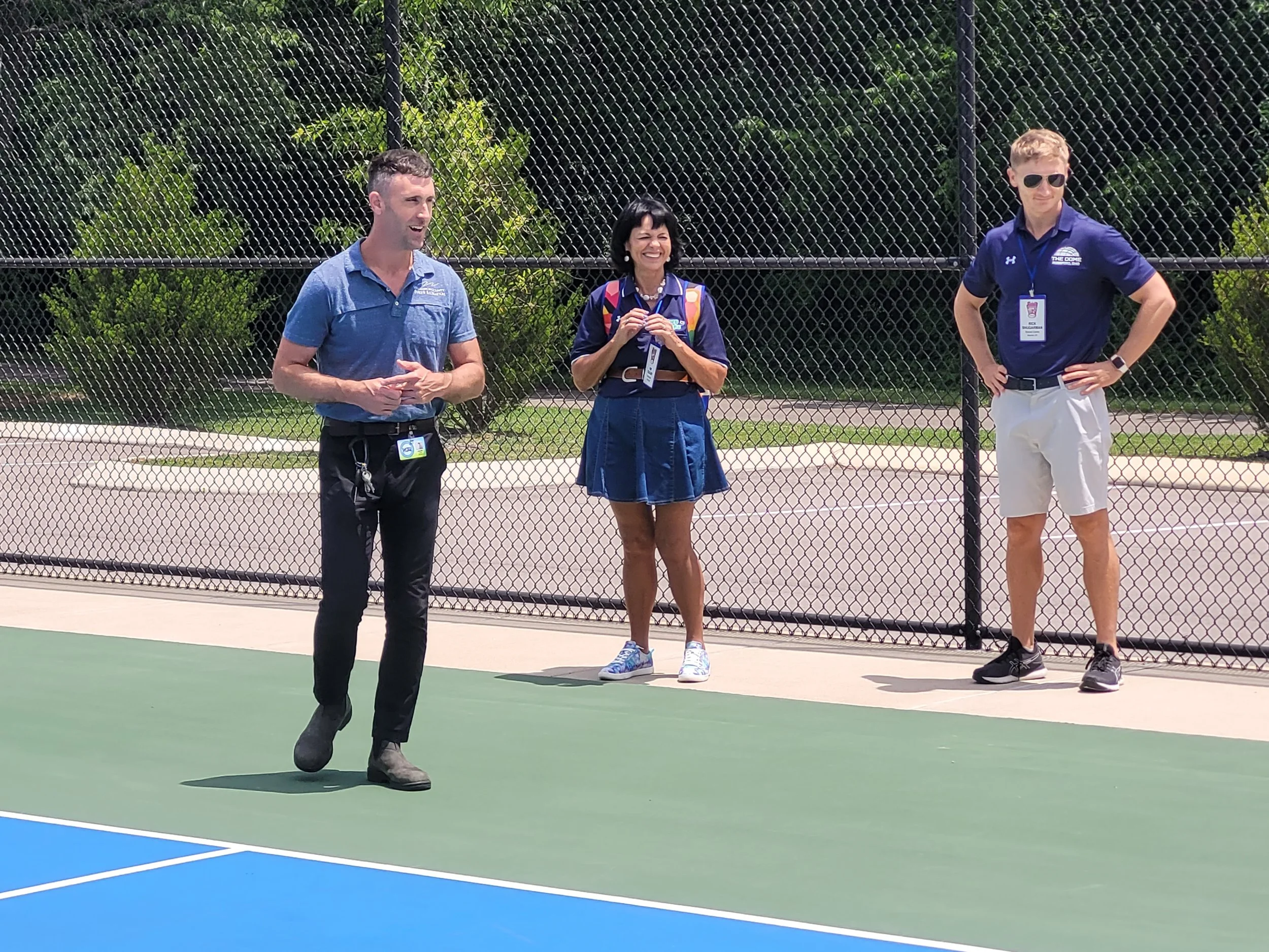 Three people standing on a tennis court behind a chain-link fence, enjoying a sunny day. The man on the left is wearing a blue shirt and black pants, the woman in the middle is smiling and wearing a blue dress with a backpack, and the man on the righ