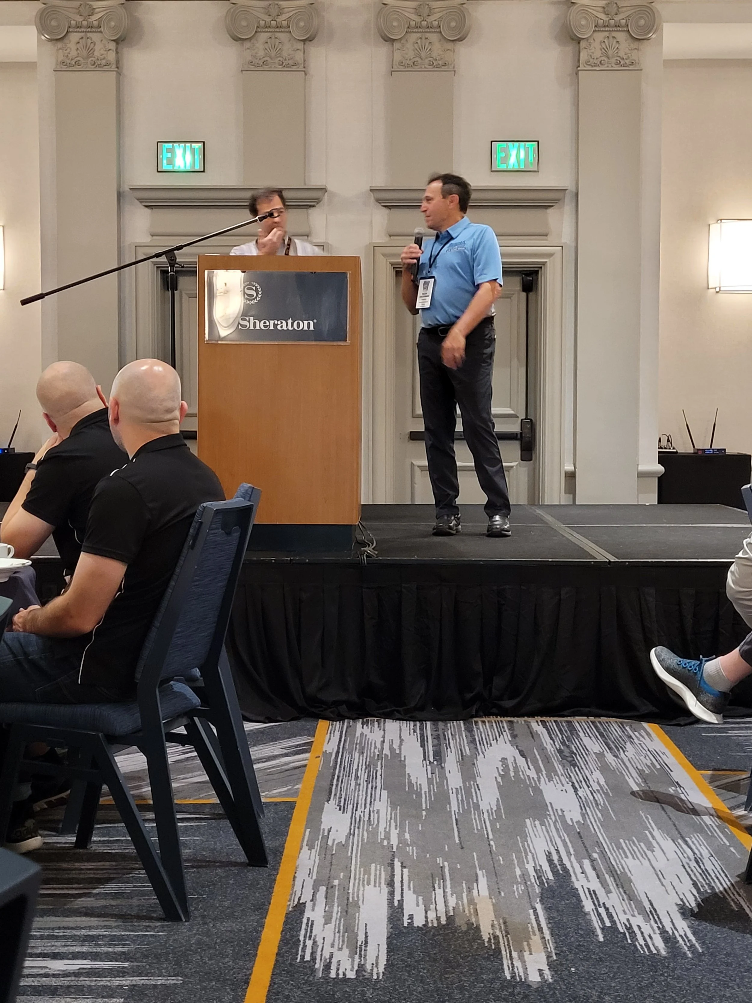 A man holding a microphone receiving a speech at a conference in a hotel ballroom with a podium displaying the Sheraton logo, and a man behind the podium. Audience members seated at tables watch the presentation.