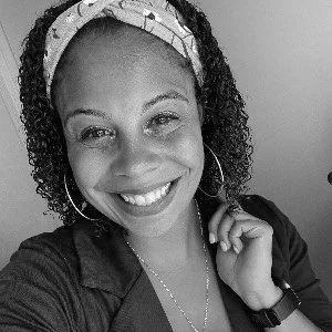 Smiling woman with curly hair, wearing hoop earrings and a headband, in black and white.