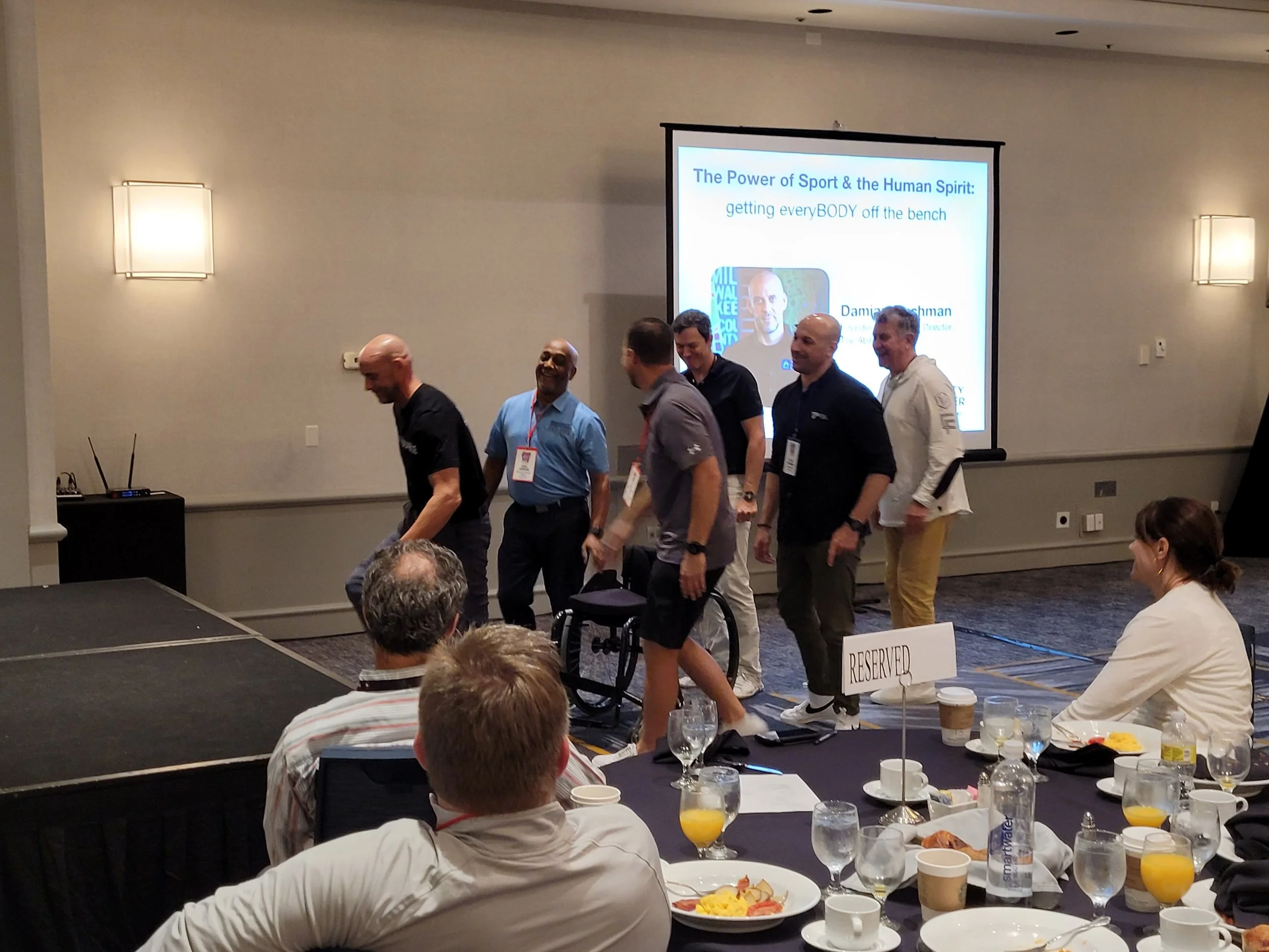 Group of conference attendees walking past a presentation screen in a large room with tables set for breakfast, filled with coffee cups, glasses of orange juice, and plates of food; reserved sign on one table; dim lighting with wall-mounted lights.