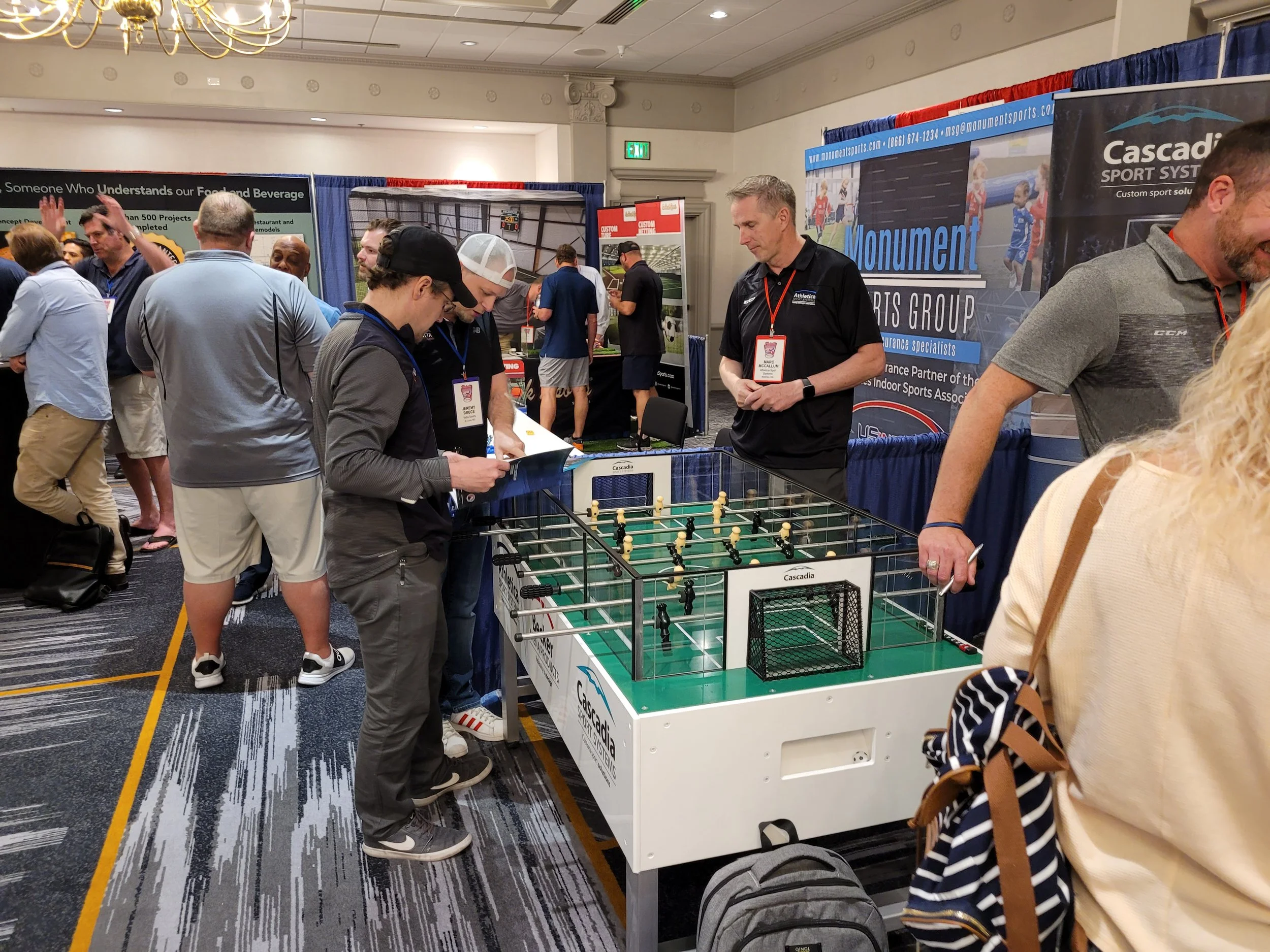 Several people attending a sports exhibition, looking at a foosball table display. There are banners with company logos in the background, and a blue curtain segment. Attendees are engaged, some examining materials on the table. The room has a patter