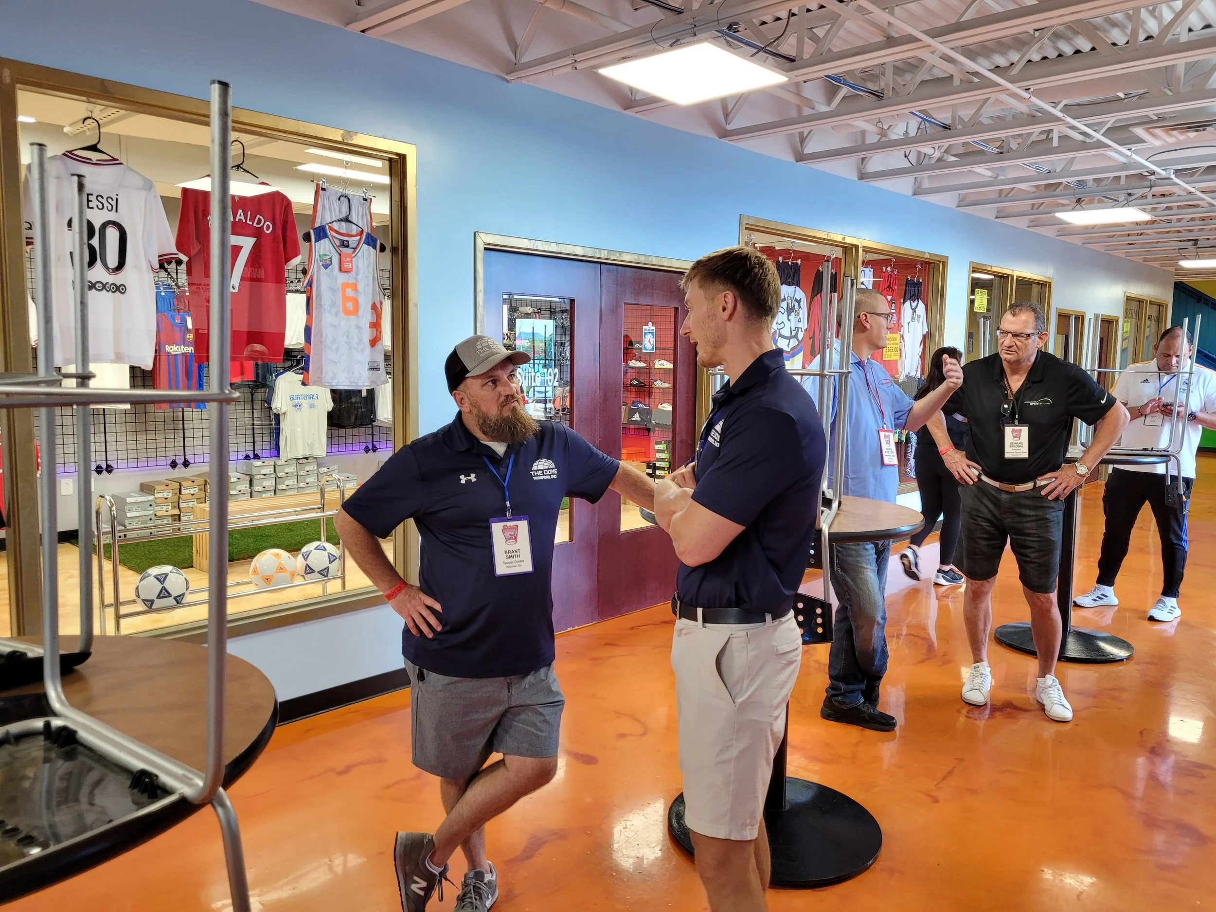 Several men are standing and talking inside a sports merchandise store, with jerseys and soccer balls displayed. Two men in the foreground are engaged in conversation, one wearing a dark shirt and the other in a sports polo. Other shoppers are browsing in the background.