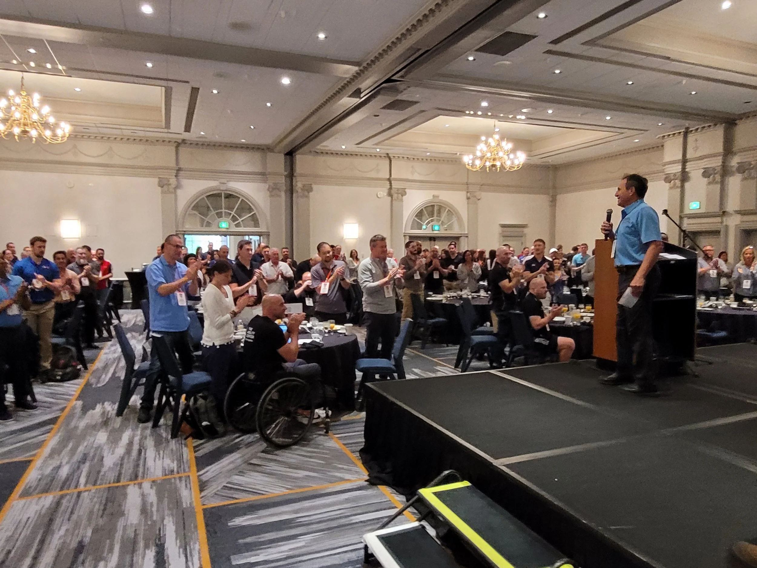 A large conference or seminar room filled with attendees standing and clapping. There is a speaker at a podium on the stage, smiling and holding a microphone. The room has high ceilings with chandeliers and decorative wall moldings.