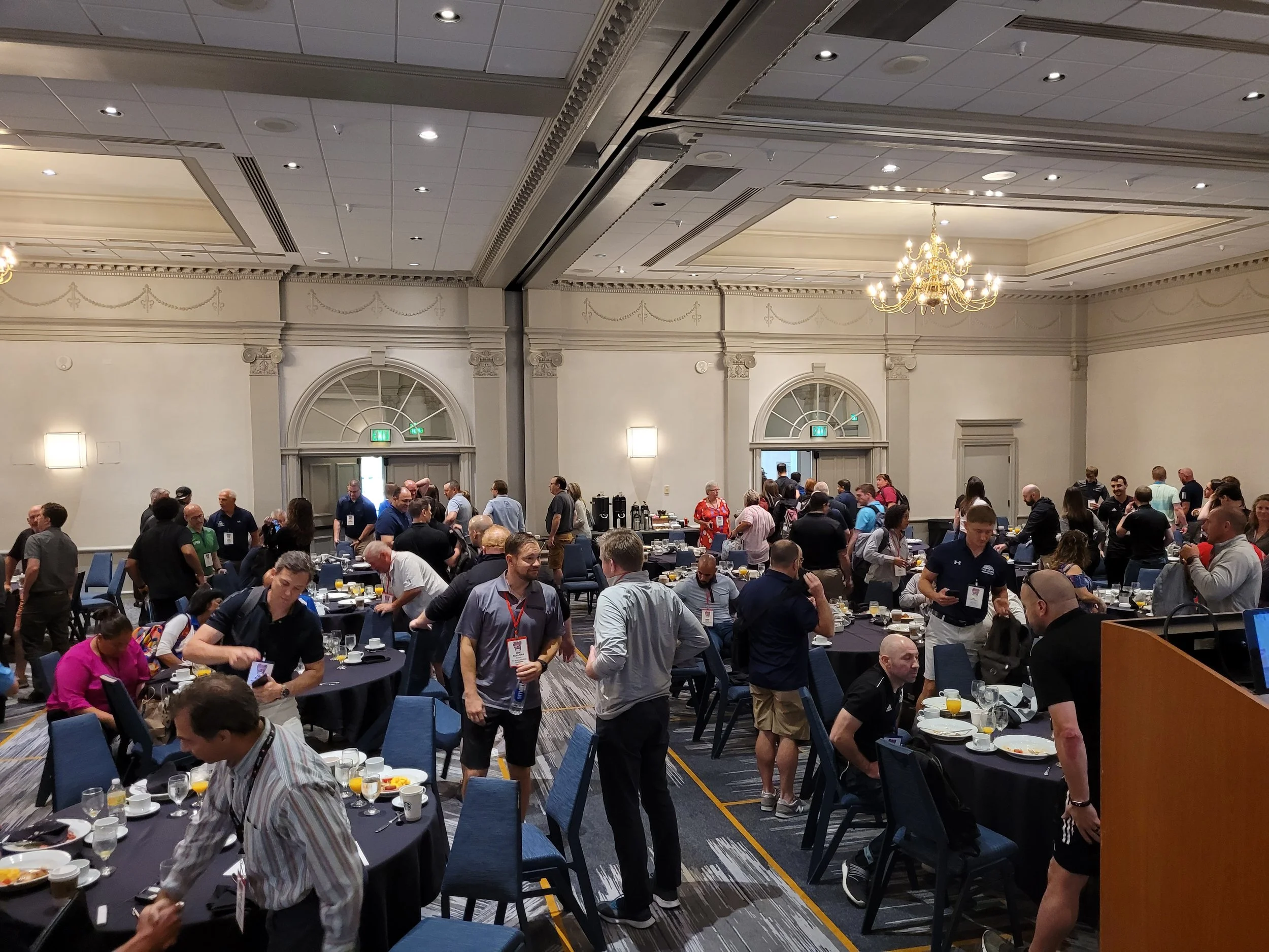 A large group of people gathered in a spacious banquet room with elegant white walls and ornate crown molding. The attendees are mingling and sitting at tables set with breakfast food and drinks, engaging in conversations. The room is decorated with 