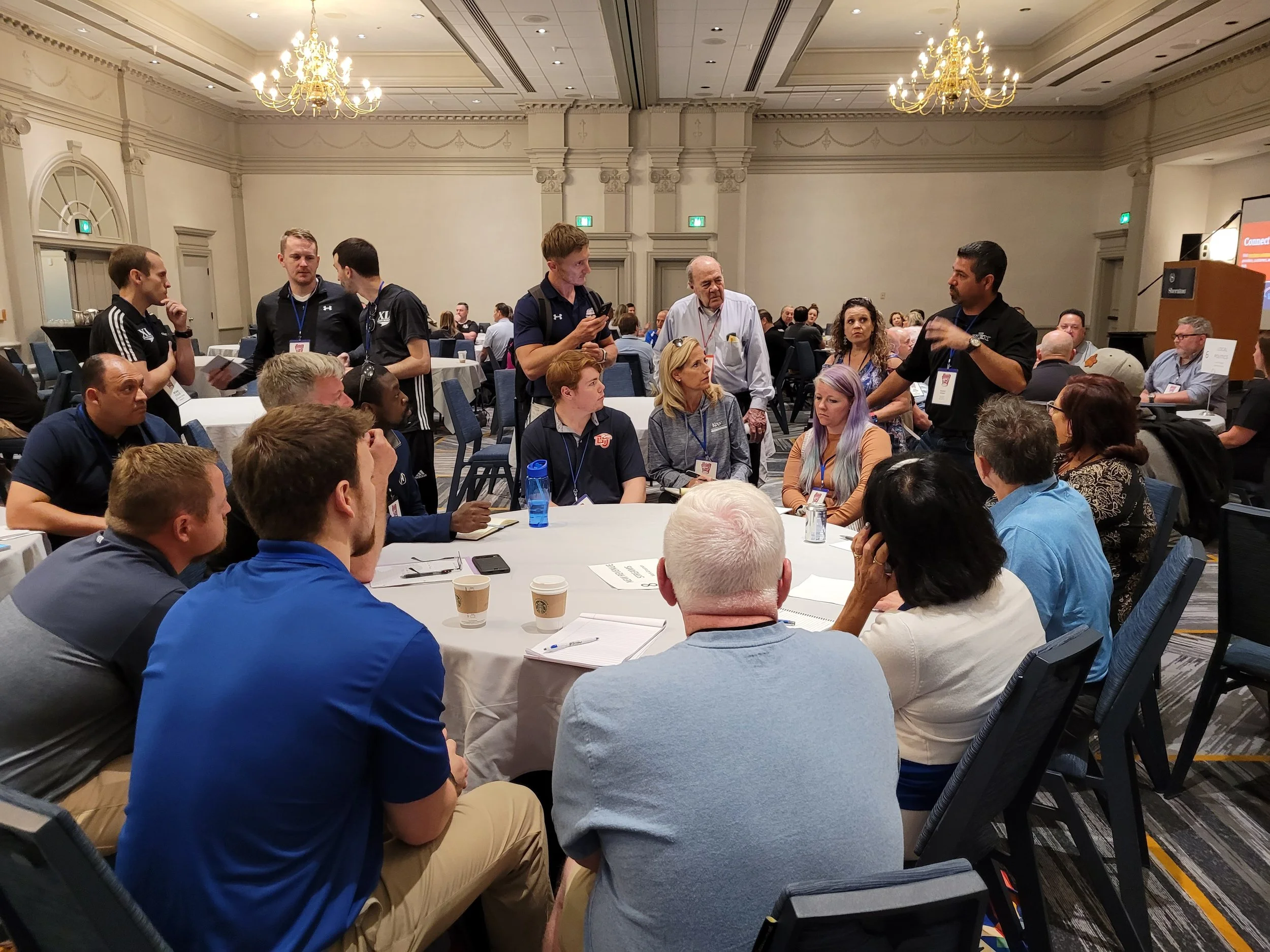 A group of people attending a conference or seminar in a large, ornately decorated room, sitting at round tables, with some standing and talking, some taking notes, and others listening attentively.