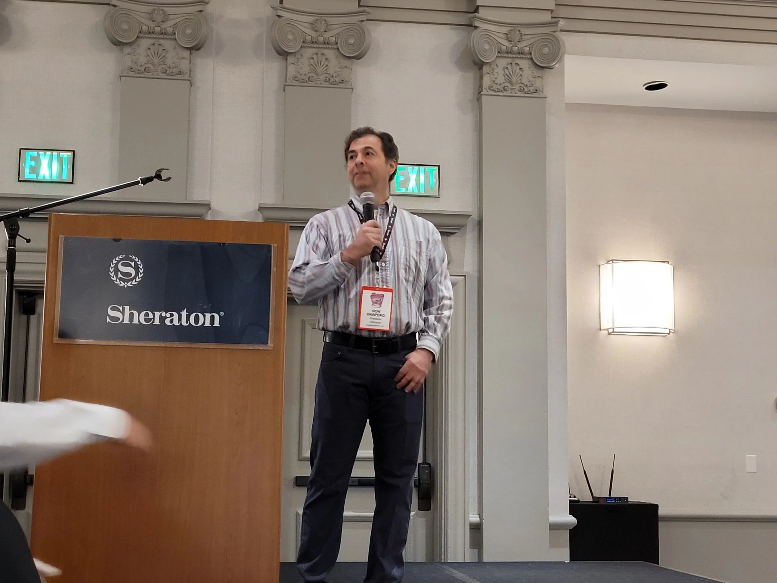 A man in a striped shirt holding a microphone stands at a podium in a conference room with decorative columns and two illuminated exit signs behind him.