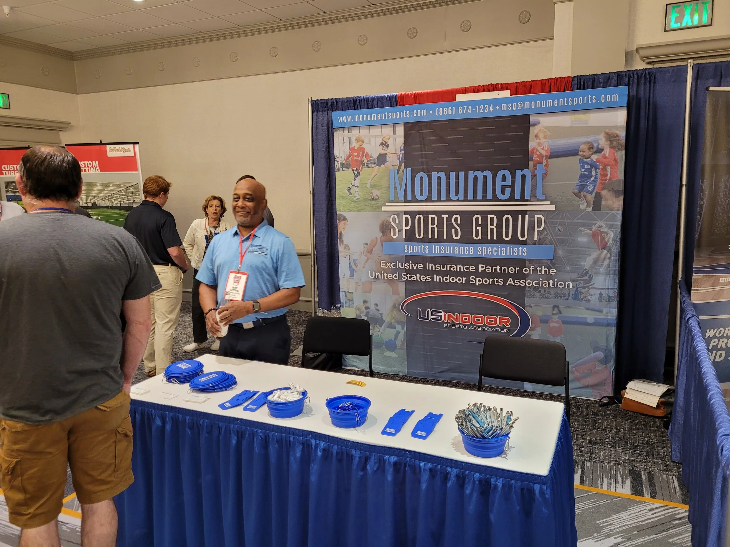 People at a trade show booth for Monument Sports Group with a large banner displaying sports images and company information. The booth has promotional items like pens and lanyards on a table.