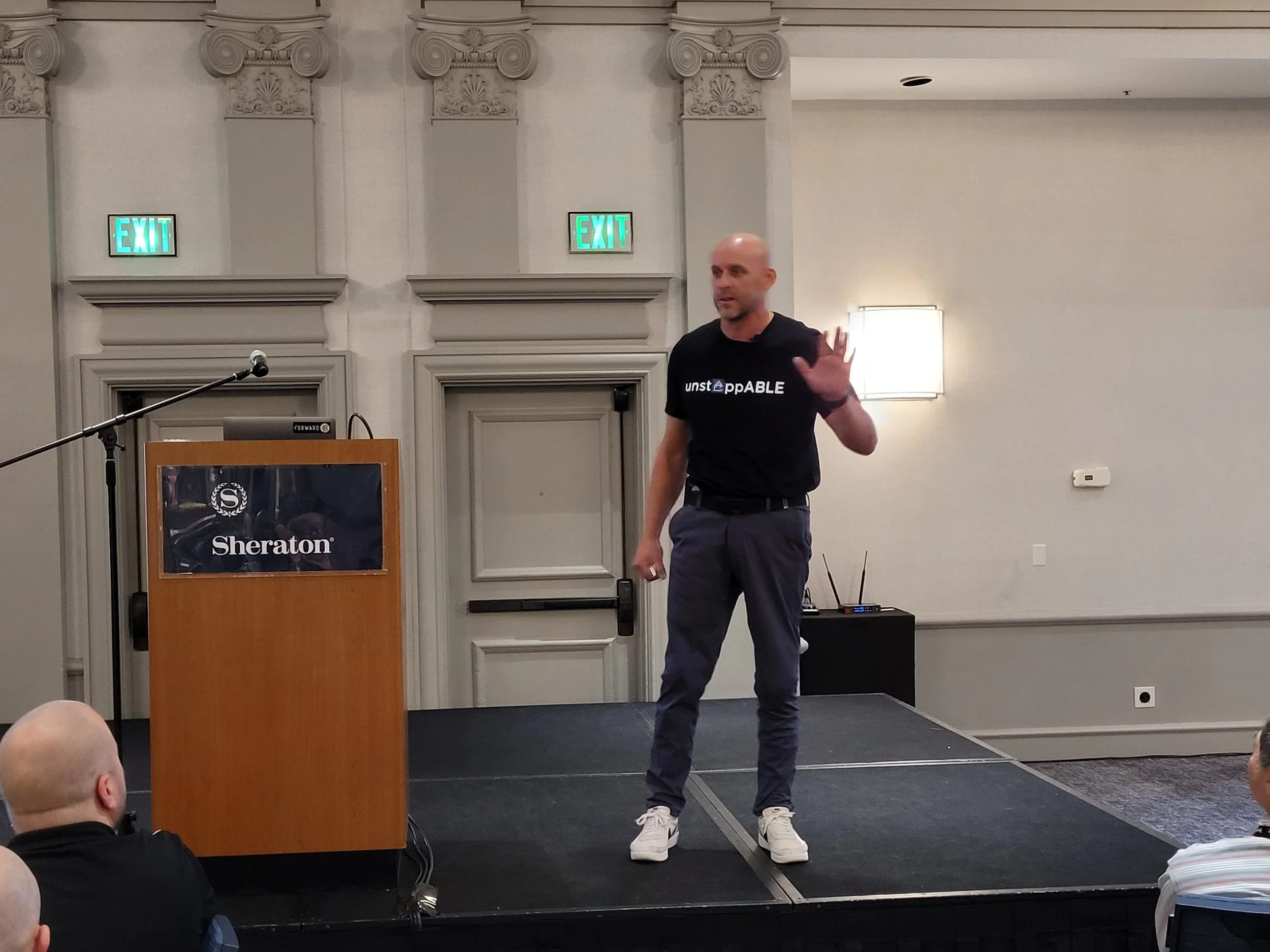 A man standing on a stage in a conference room giving a presentation. He is bald, wearing a black t-shirt with the words 'unstoppable' printed on it, and gray pants. There is a podium with a logo and a microphone on top of it to his left, and the bac