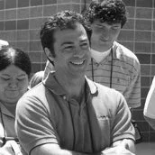 A group of people smiling outdoors near a fence, including a man with crossed arms in the foreground.