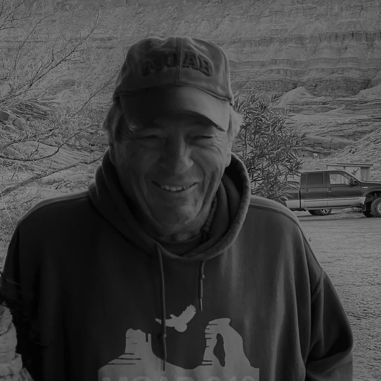 A man smiling outdoors wearing a cap, hoodie, and jacket, with a background featuring desert landscape and a truck.