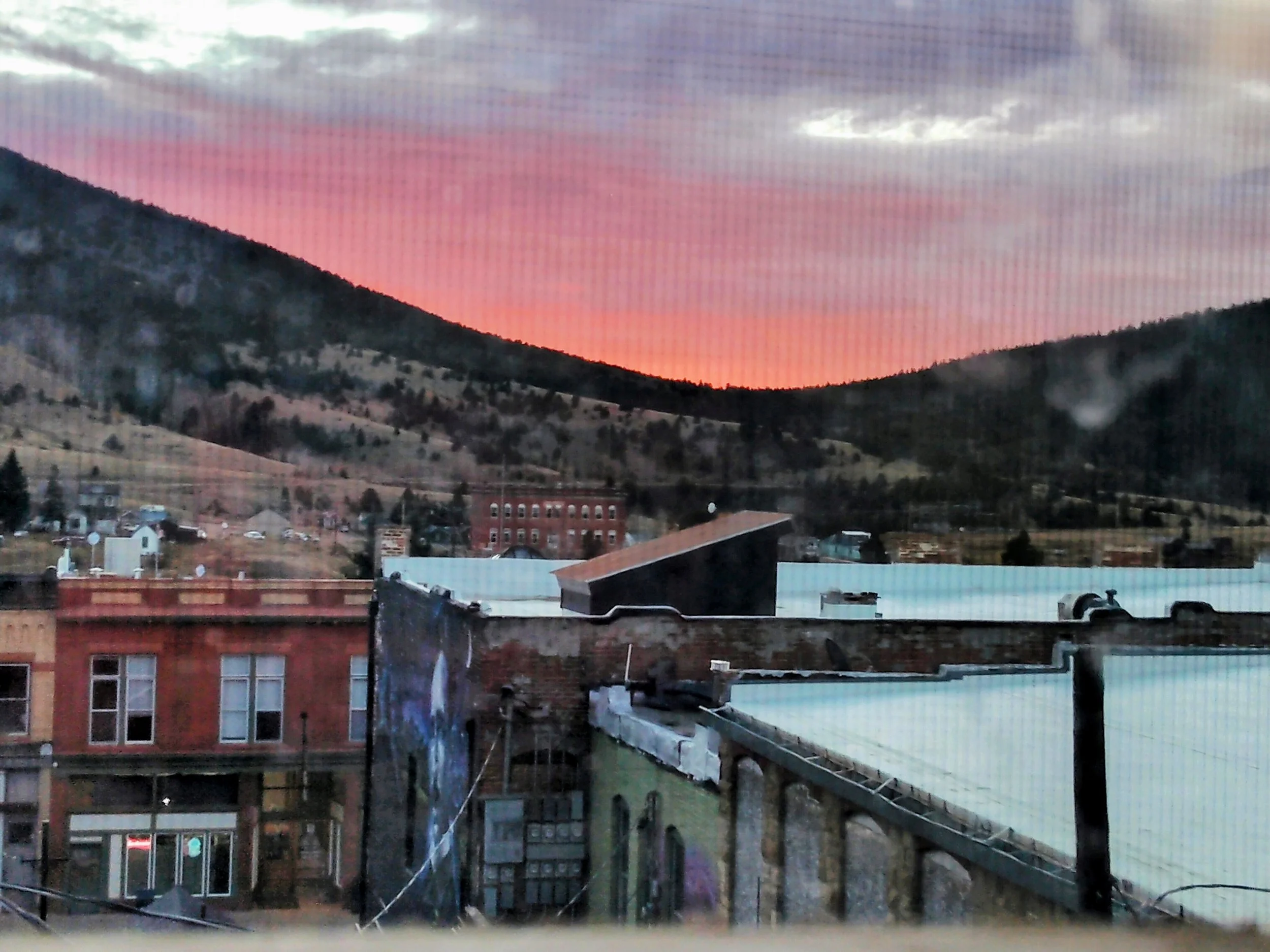 A scenic view of a sunset sky with pink and orange hues over a hillside, with a small town in the foreground consisting of buildings, rooftops, and street infrastructure.