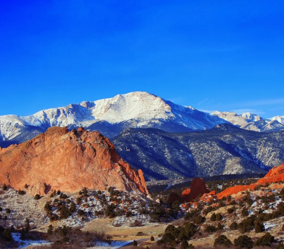Snow-capped mountains with red sandstone formations in foreground and a clear blue sky.