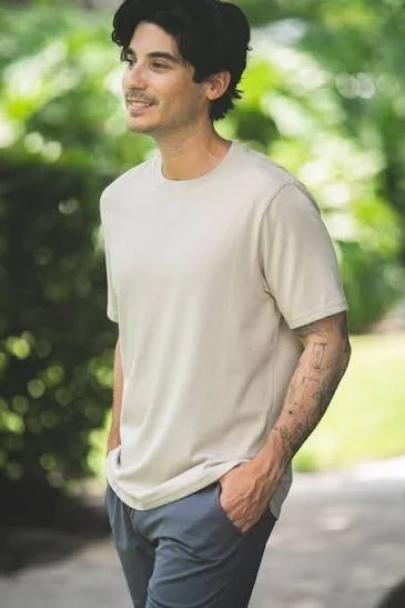 A young man with dark hair, smiling, walking outdoors on a sunny day, wearing a white t-shirt and blue pants, with tattoos on his left arm.