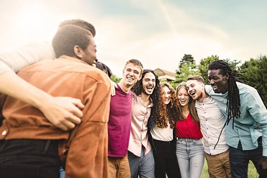 A group of seven diverse young adults standing together outdoors, smiling and laughing as they enjoy each other's company on a bright day.