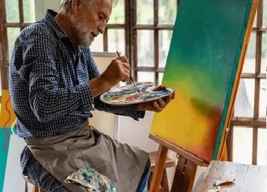 An elderly man with white hair and a beard painting on a colorful canvas in a bright room with large windows.